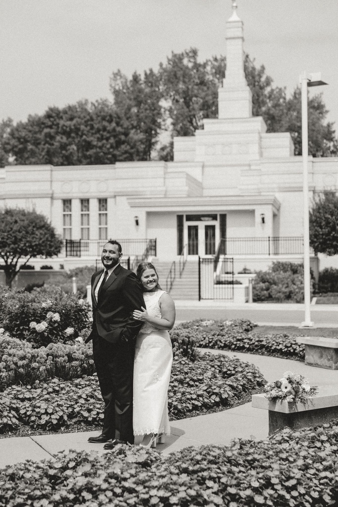 Black and white photo of a smiling couple in wedding attire posing in front of an LDS temple garden.