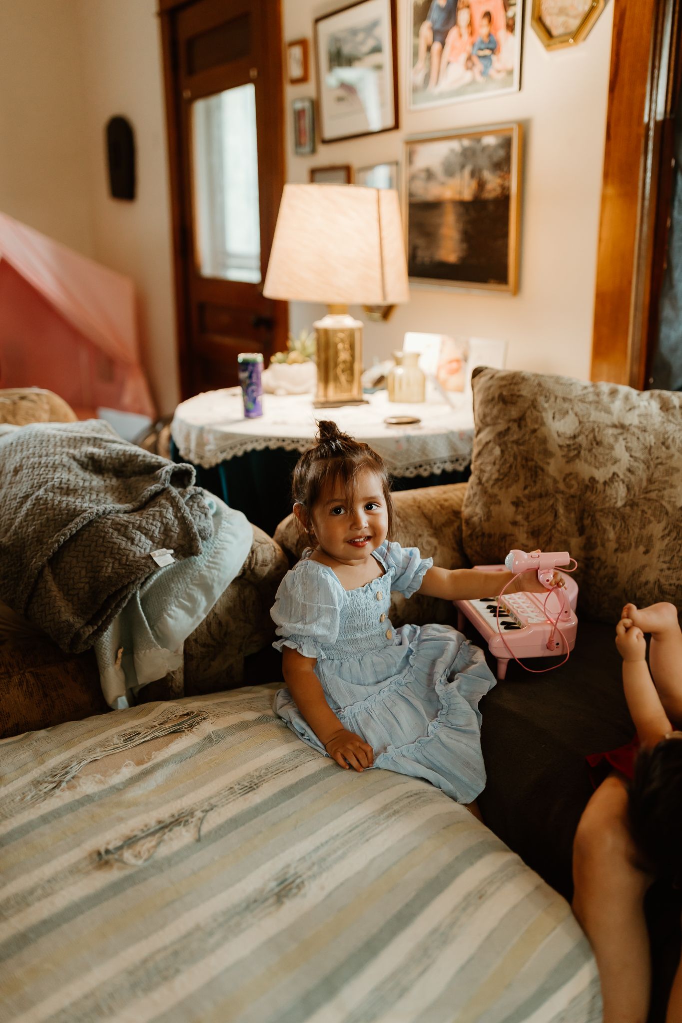 A young girl in a blue dress sits smiling on a couch beside a pink toy piano in a cozy, warmly lit living room.
