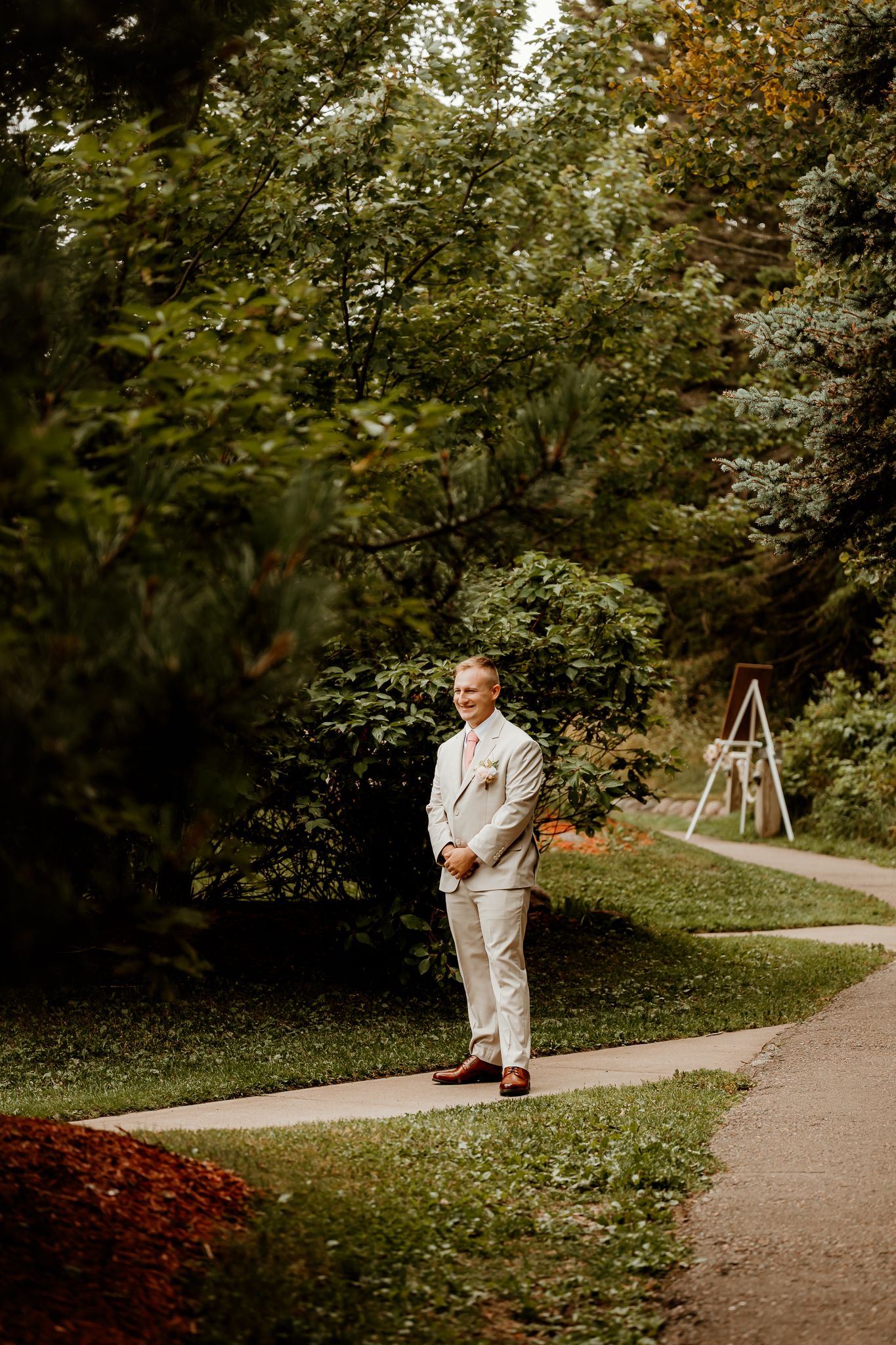 A smiling groom in a white suit stands on a garden path surrounded by lush green trees on his wedding day.