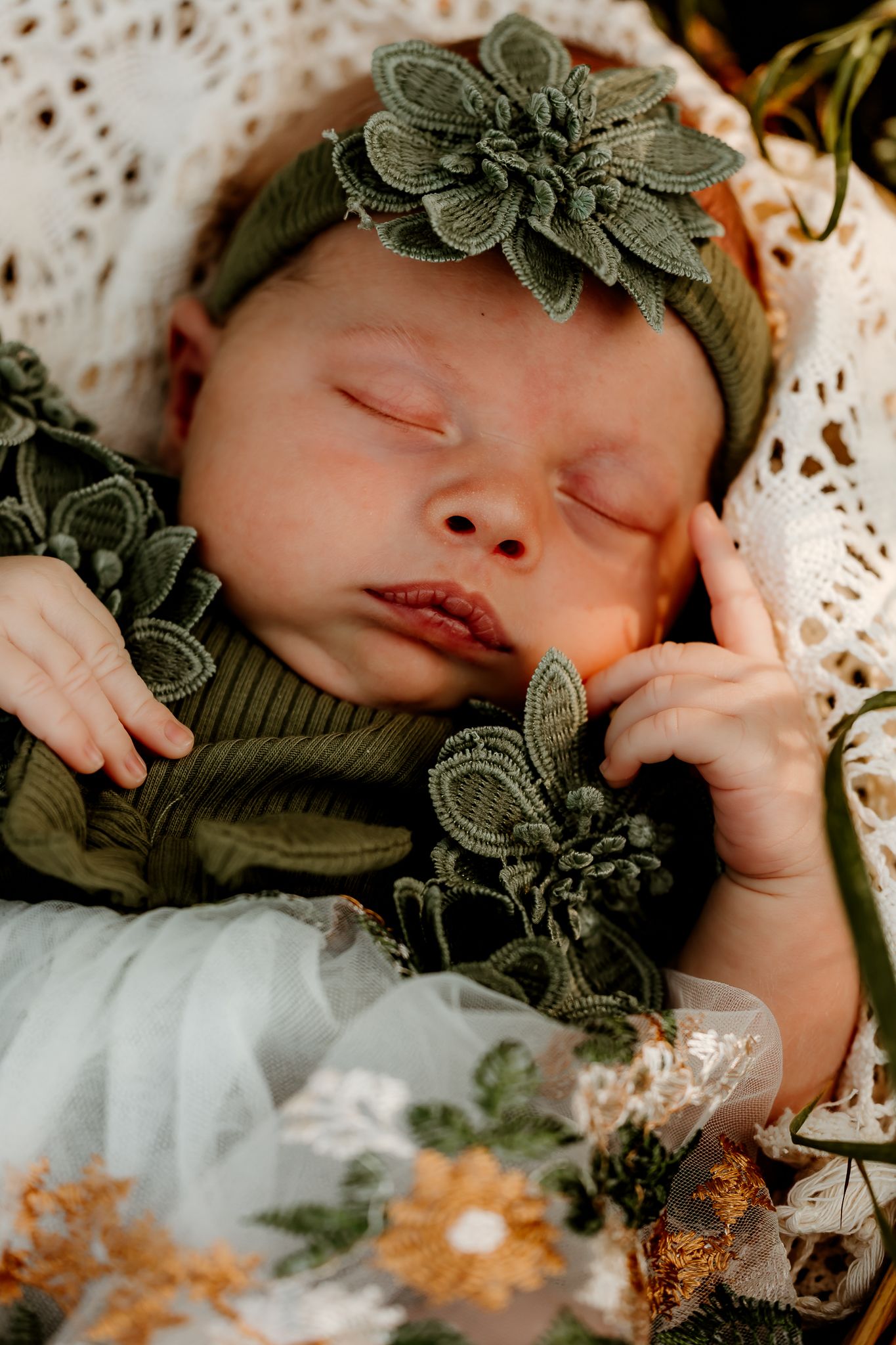 Sleeping newborn baby wearing a green ribbed outfit and floral headband, surrounded by delicate flowers and lace.
