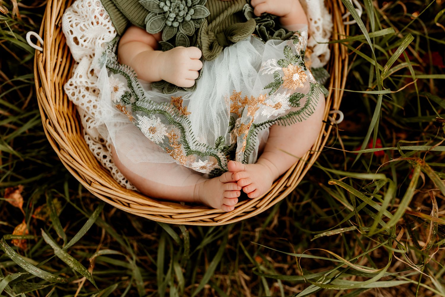 A baby in a floral green dress rests in a wicker basket surrounded by lush green grass.