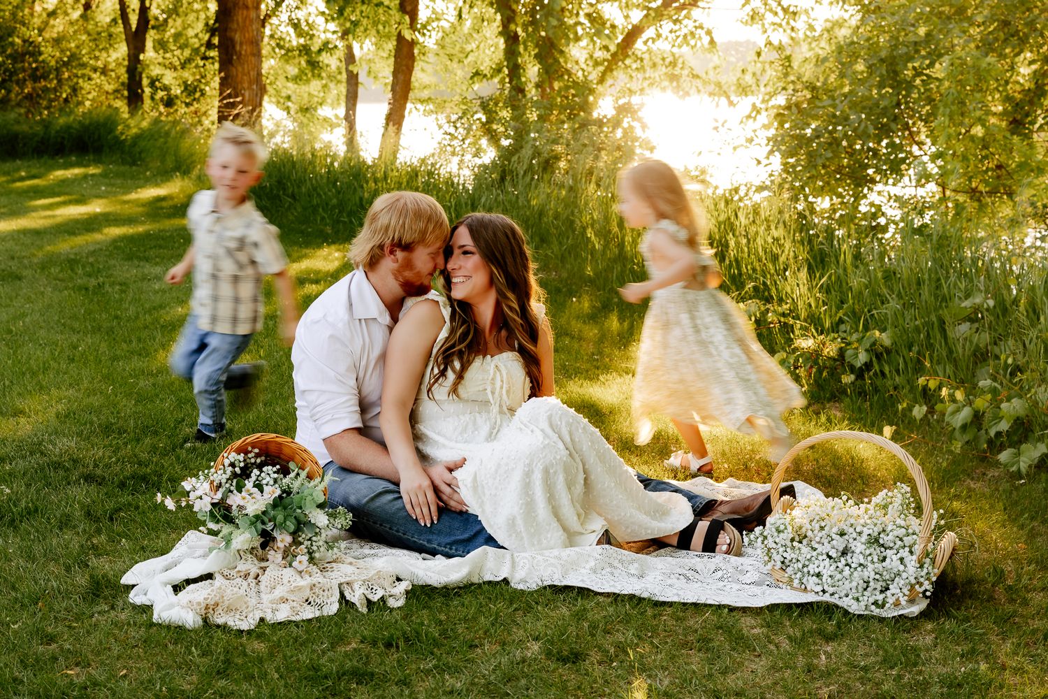 Couple sharing a kiss on a picnic blanket with flowers while children play in golden sunlight.