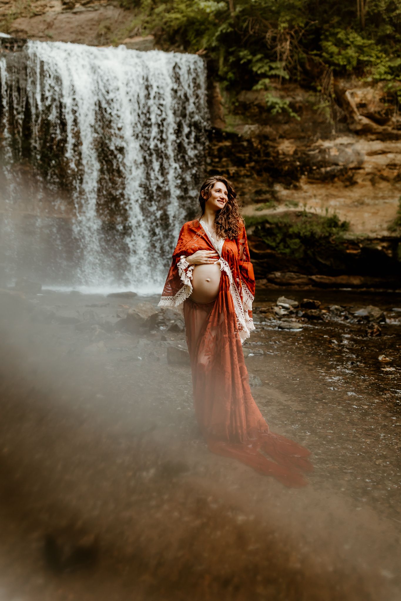 Pregnant woman in rust orange gown stands before a waterfall, cradling her baby bump in a serene outdoor setting.
