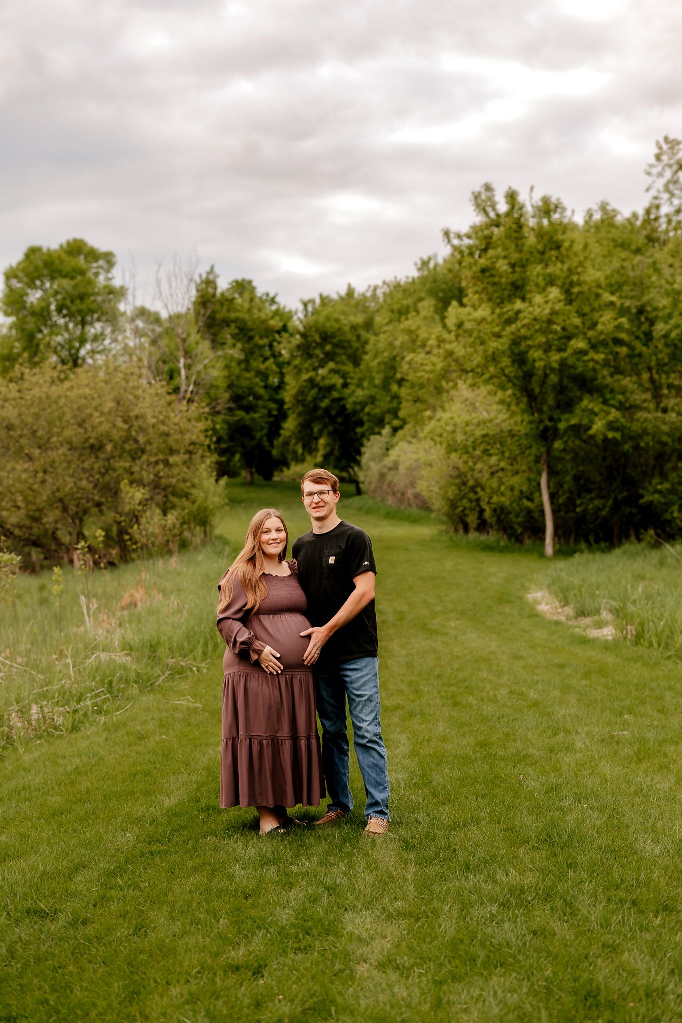 A smiling couple poses on a green grass path, gently holding the woman's pregnant belly outdoors.