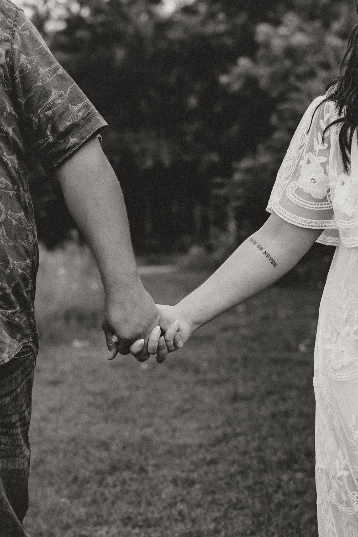 Black and white close-up of couple holding hands, woman with arm tattoo in white lace dress.