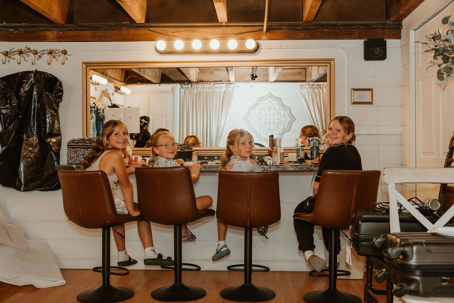 Women and children sitting at a brown leather bar counter inside a bright, rustic salon or event space.