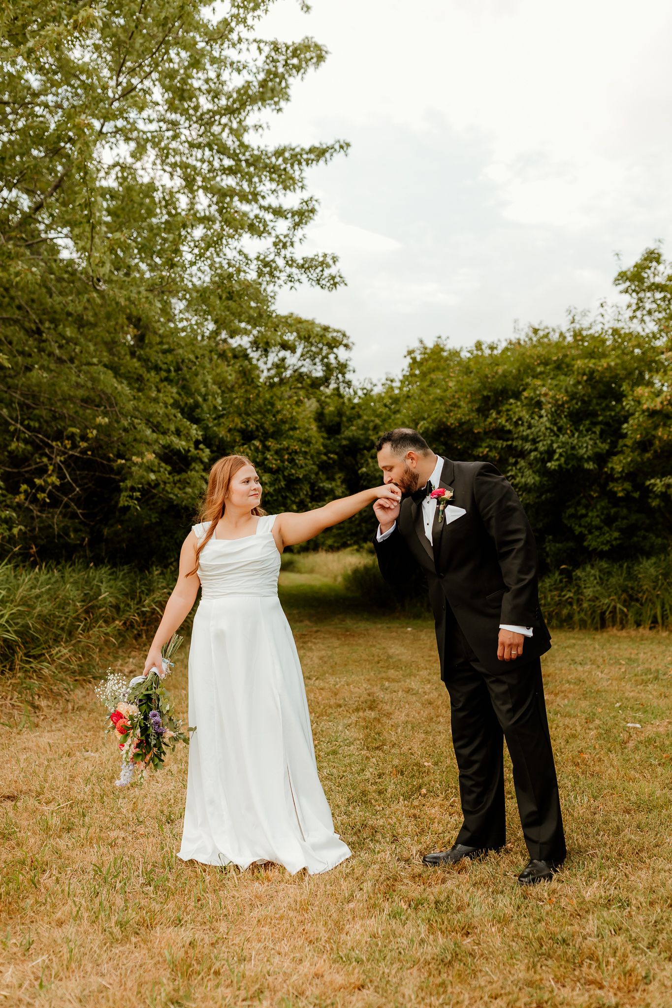 Groom in black tuxedo kisses bride's hand as she smiles in white gown, holding bouquet in green field.