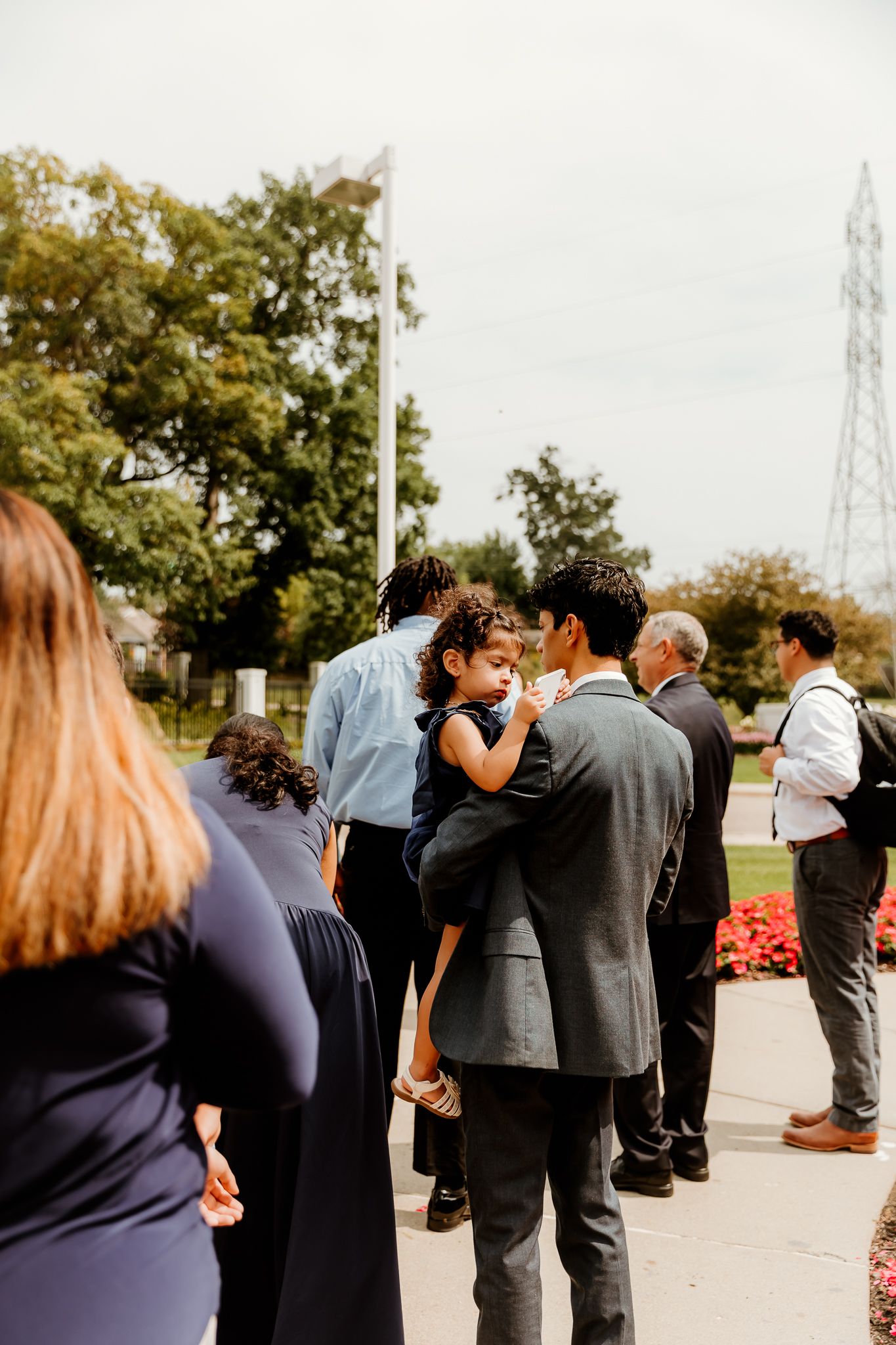 Group of elegantly dressed guests gathering outdoors at a formal event, with a man holding a young child.