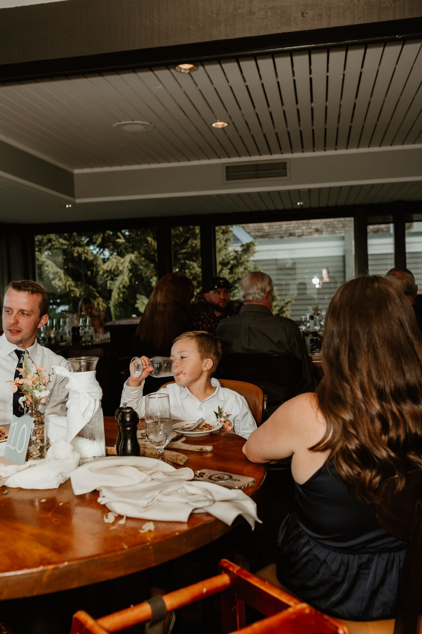 Guests smile and drink at a wooden table during a wedding reception in a warmly lit restaurant venue.