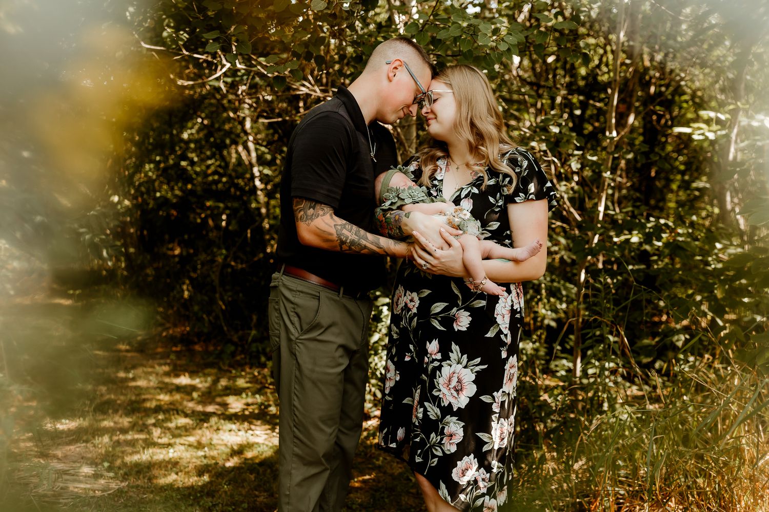 A couple embraces tenderly in a sunlit forest, the woman in a floral dress, the man with tattoos, touching foreheads romantically.