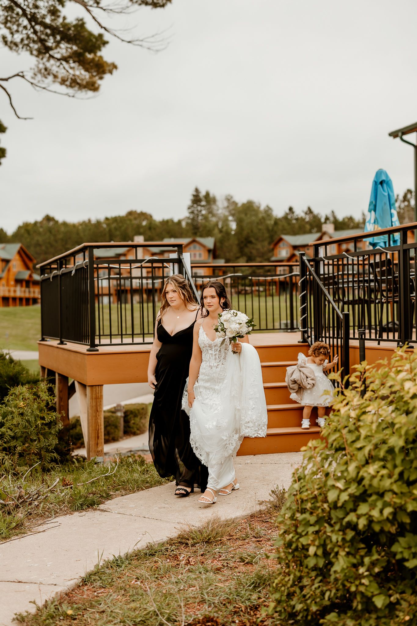 Bride in a lace wedding gown holding a white bouquet walks beside a bridesmaid in a black dress near a wooden deck and a flower girl.