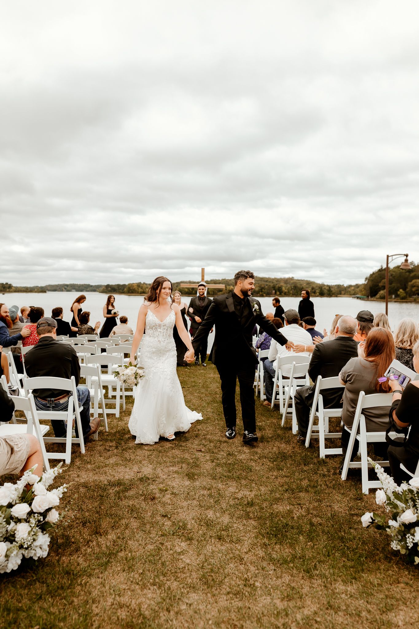 Newlyweds hold hands and walk down the aisle between white chairs of smiling guests at an outdoor lakeside wedding ceremony.