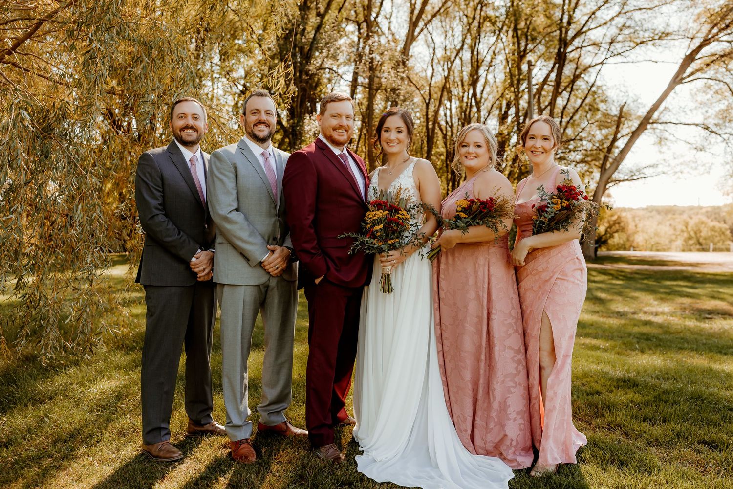 Wedding party of three groomsmen, a bride in a white gown, and two bridesmaids in pink dresses holding bouquets under sunlit trees.