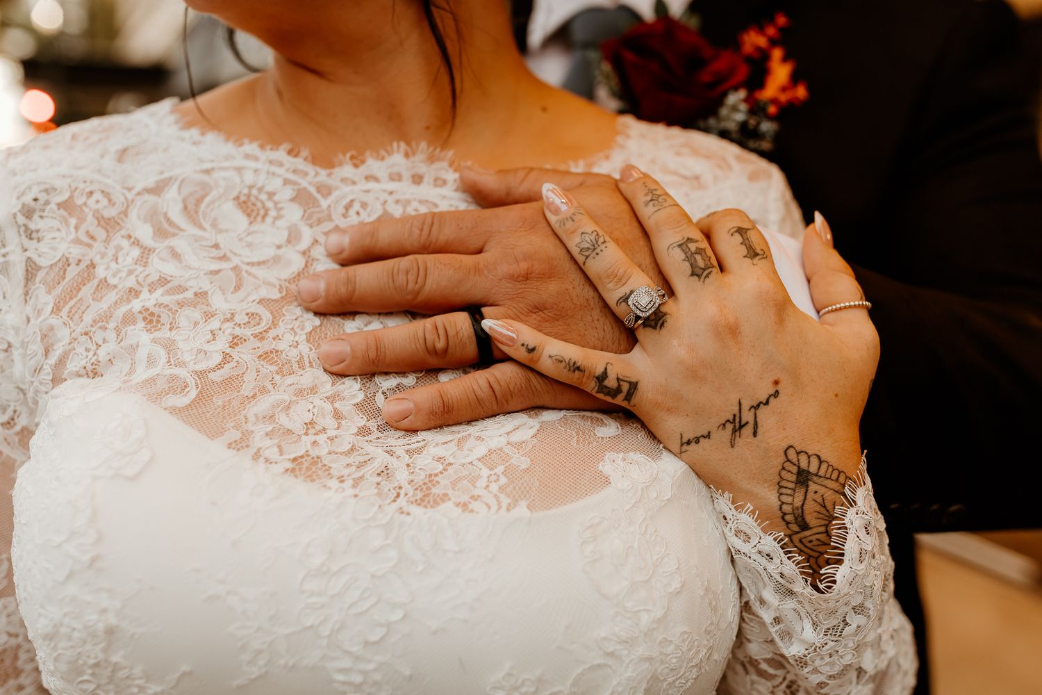 Bride in a white lace wedding dress with a tattooed hand and silver ring resting over a partner's hand in a dark suit.