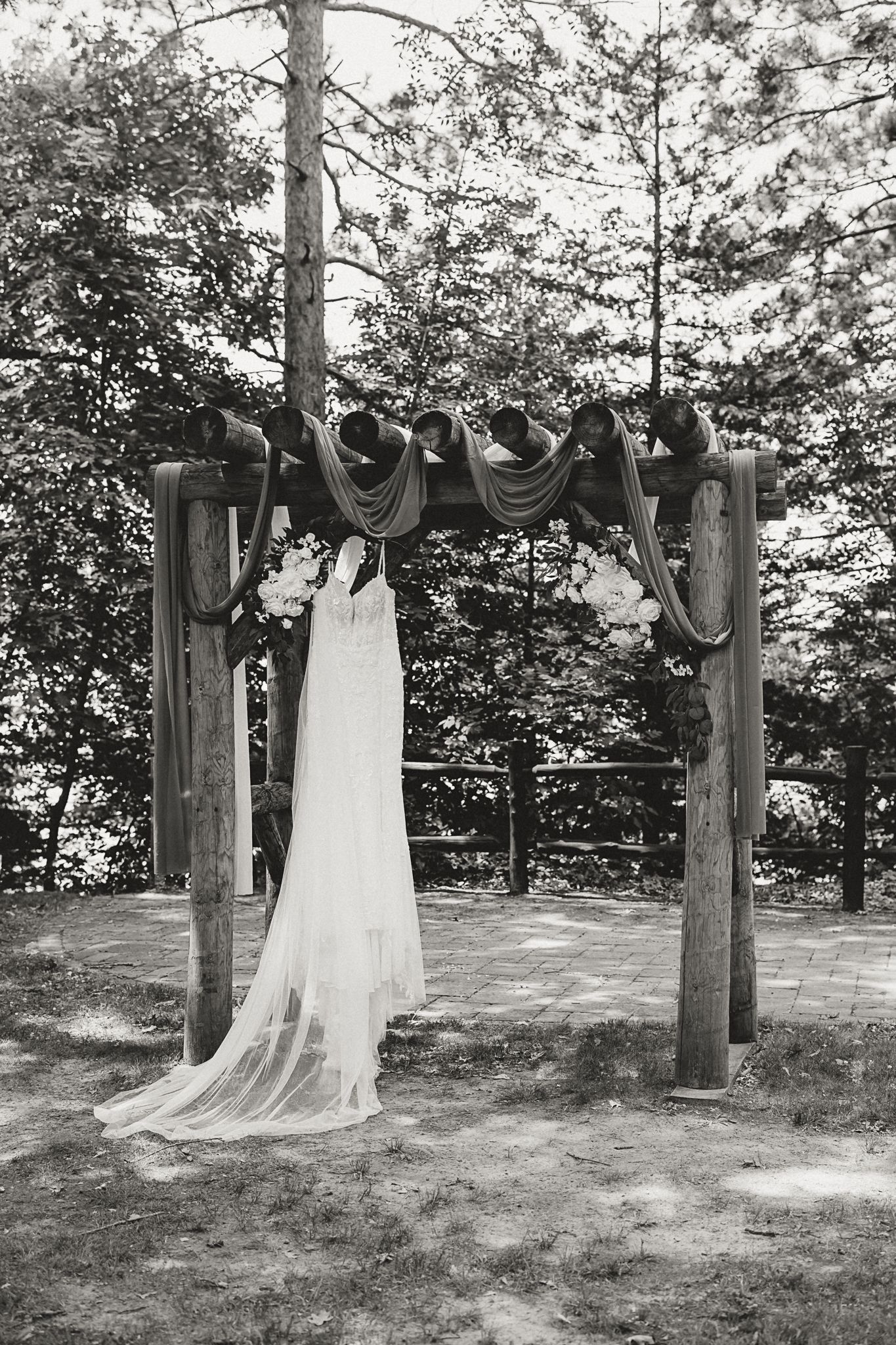 Black and white photo of a wedding dress hanging on a rustic wooden arch decorated with fabric and flowers outdoors.