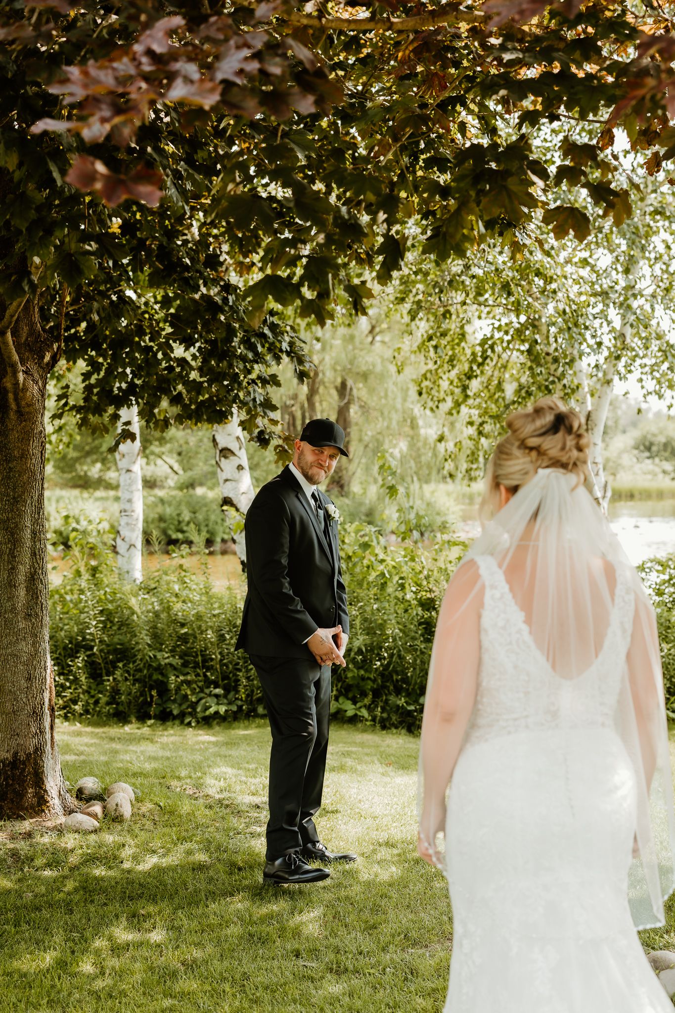Bride in white lace gown with veil approaches groom in black suit for a romantic first look outdoors.