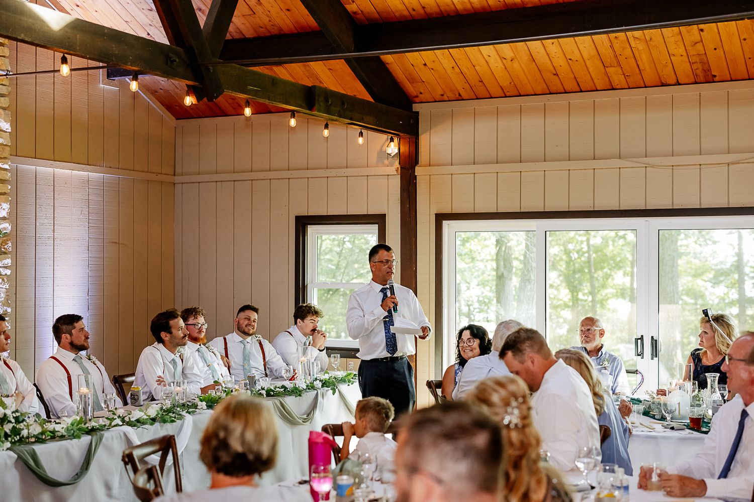 Man giving a speech at an elegant wedding reception with guests seated at decorated tables in a rustic venue.