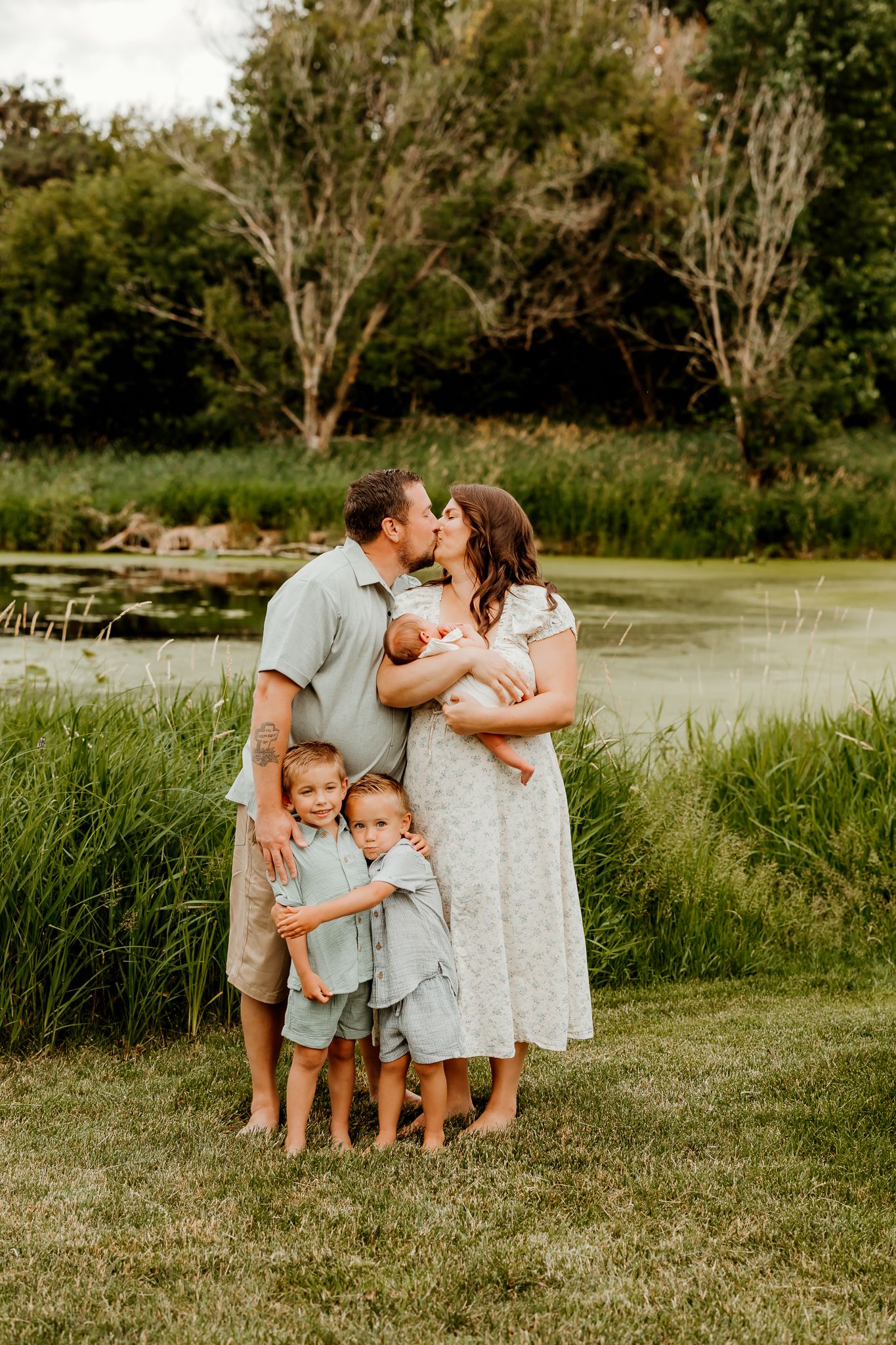 A family of five poses by a pond, parents kissing while holding a newborn, two young children hugging in front.