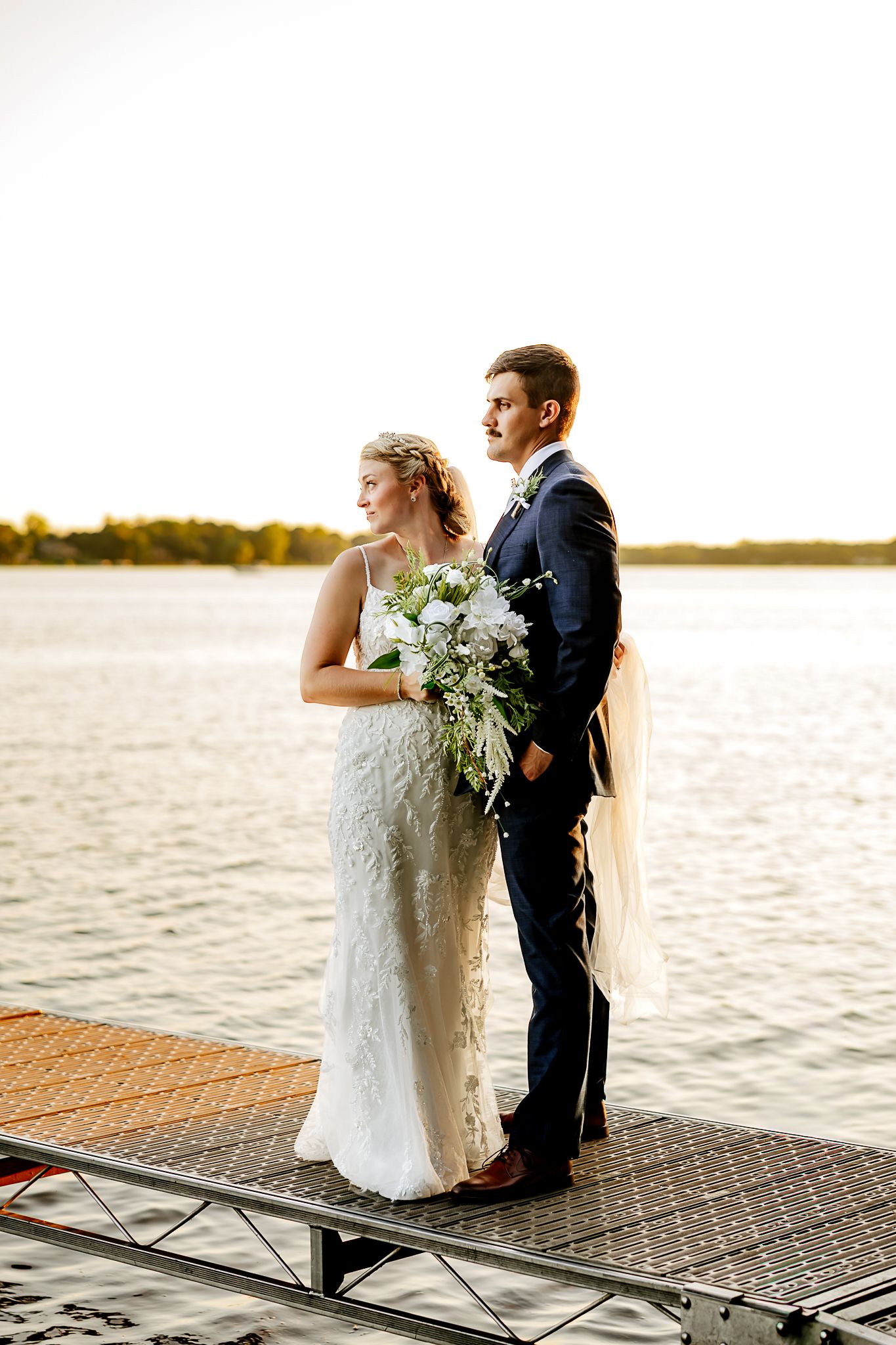 Bride and groom stand on a lakeside dock at sunset, she holds a white bouquet in a lace gown, he wears a navy suit.
