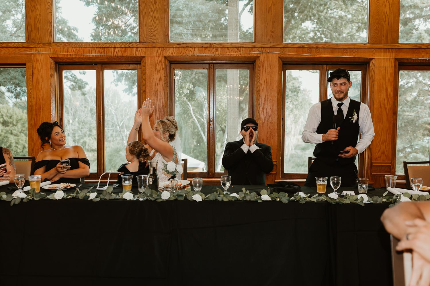 Wedding reception head table with guests, groom in vest reading speech, large windows with greenery backdrop.