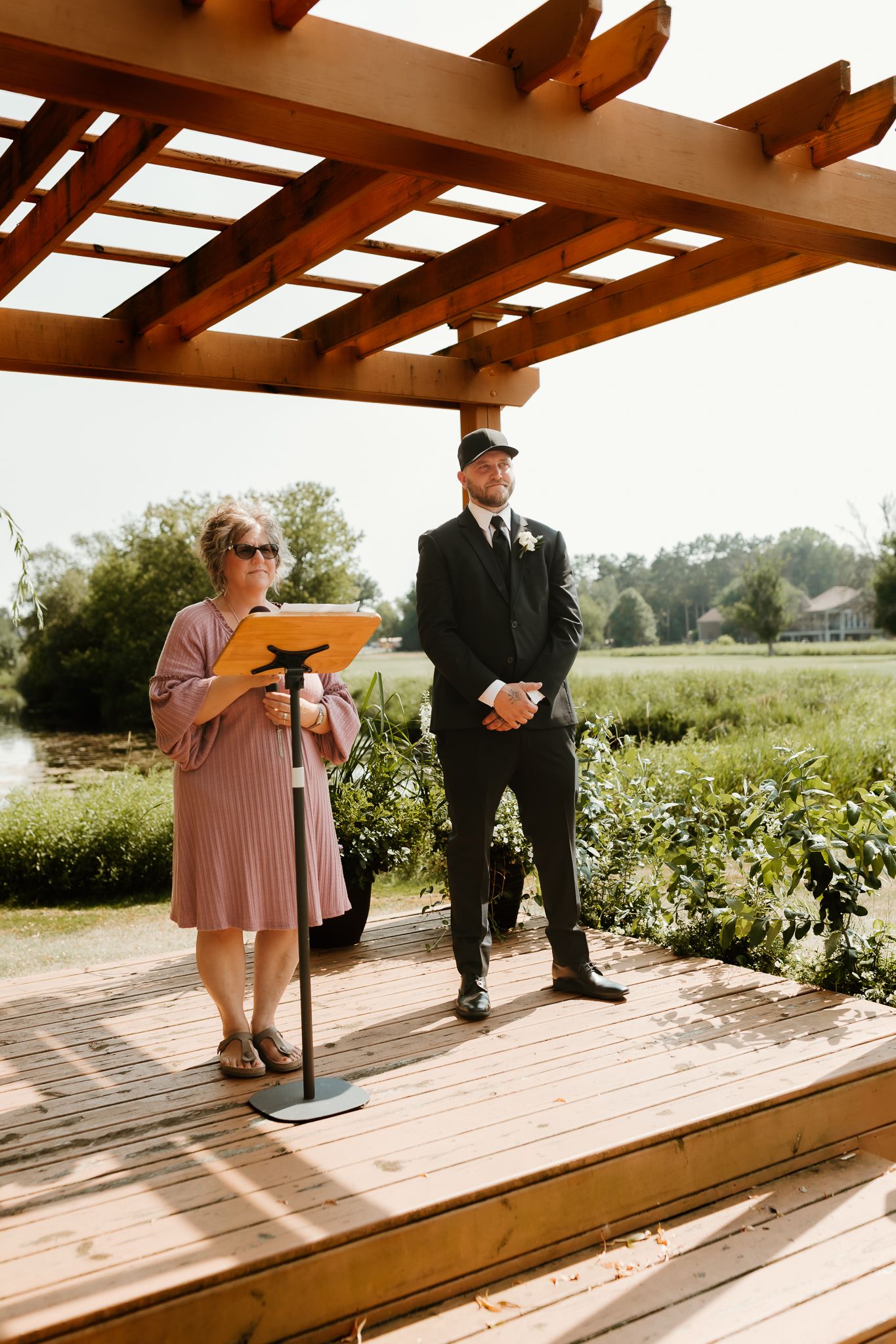 Outdoor wedding ceremony on wooden deck pergola with officiant reading and groom standing by waterfront.