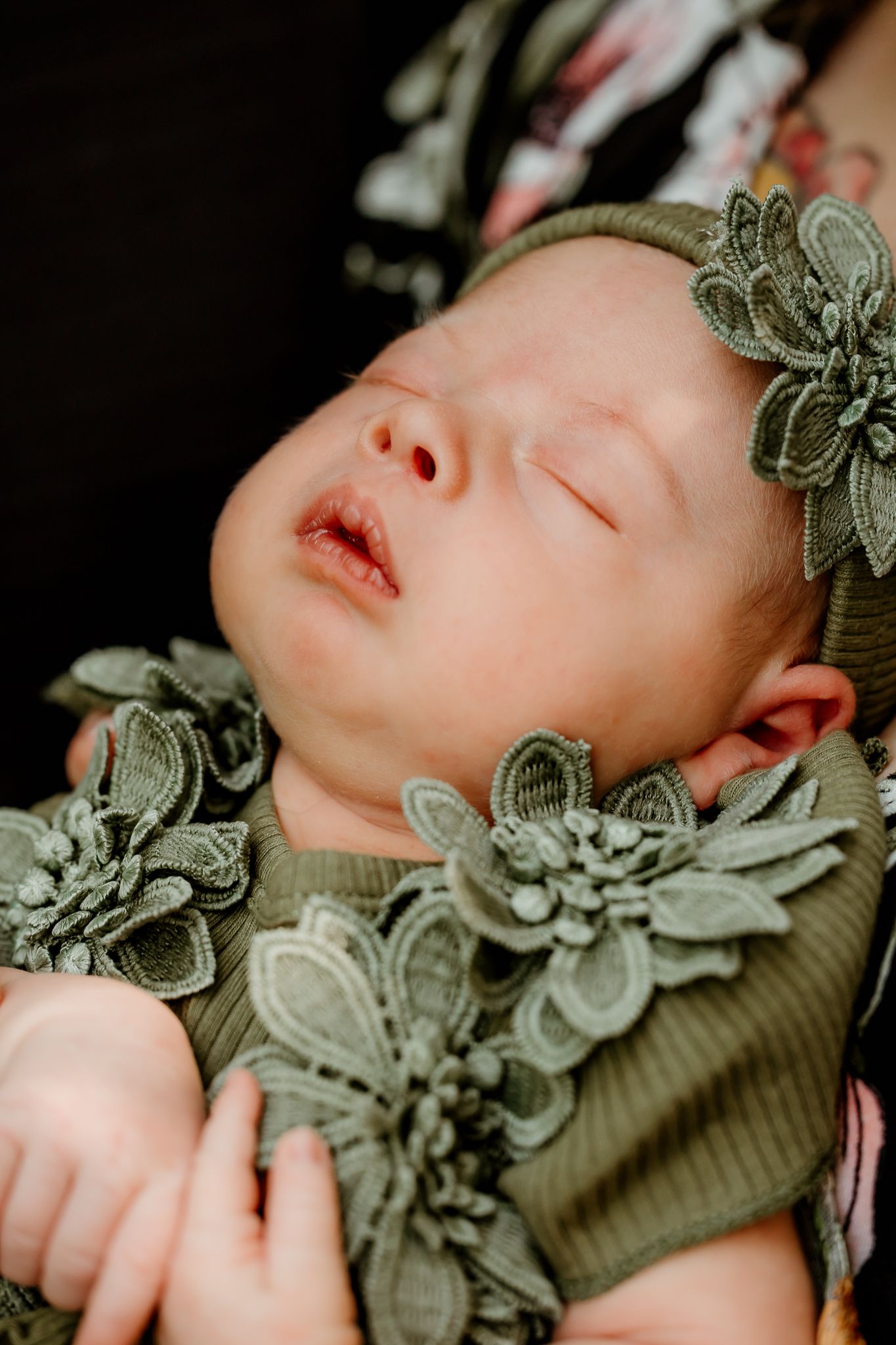 Sleeping newborn baby in olive green floral outfit and matching headband, cradled in arms.