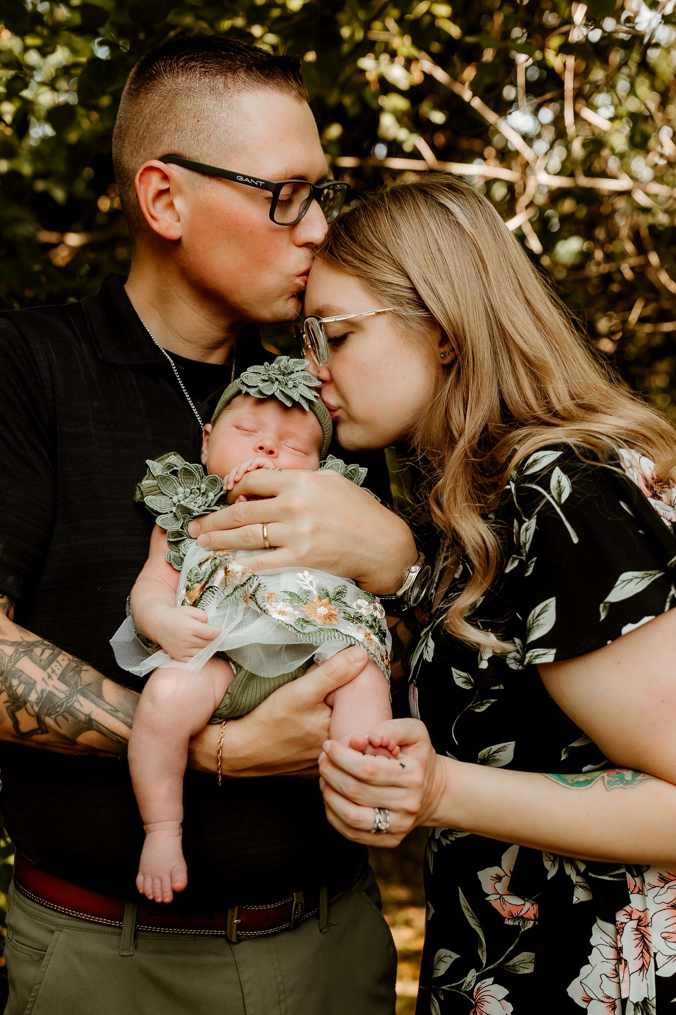 Two parents kissing their newborn baby dressed in floral outfit outdoors among green foliage.
