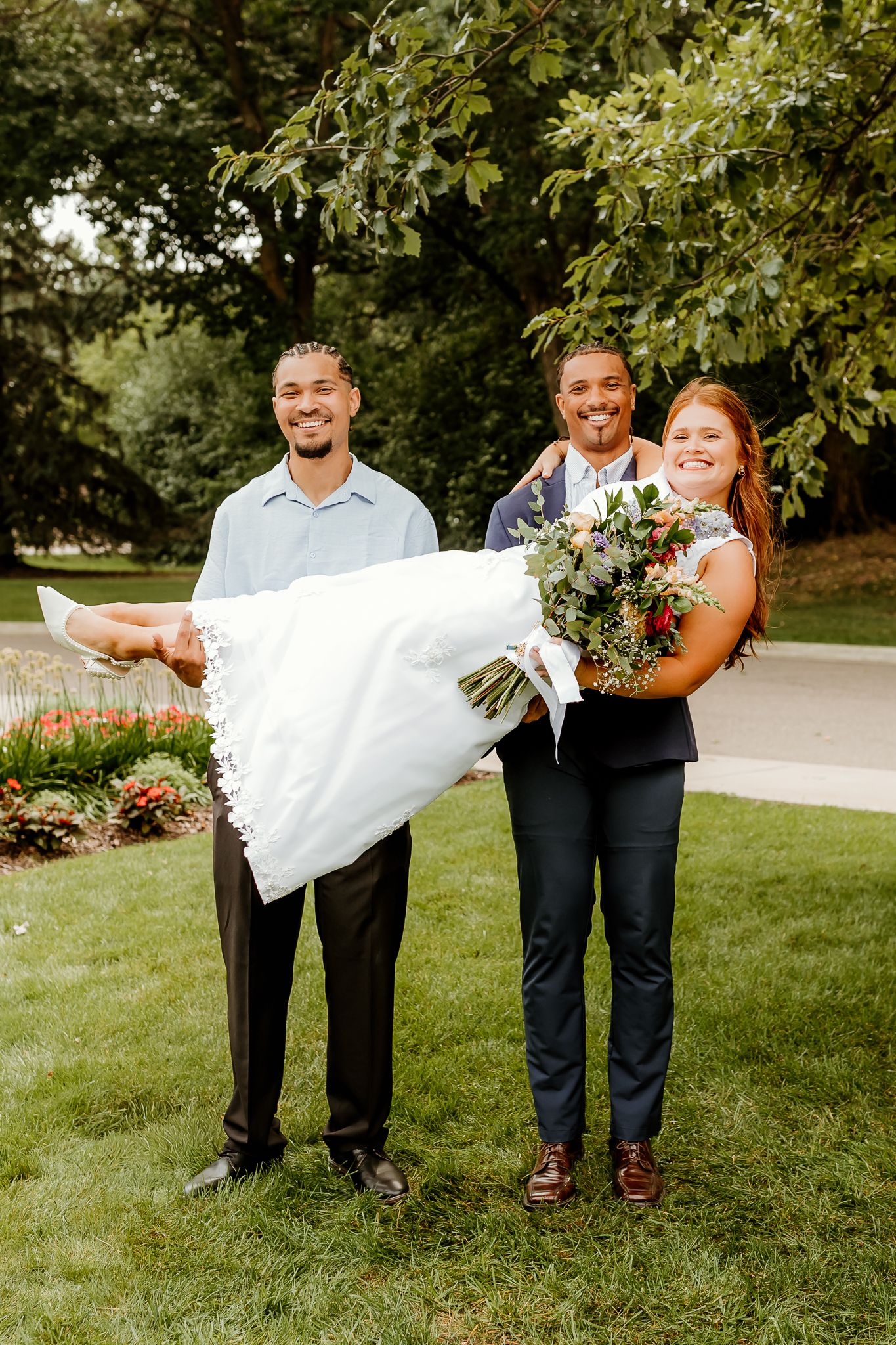 Groom carries bride in wedding dress while groomsman and bridesmaid smile outdoors in lush green garden setting.