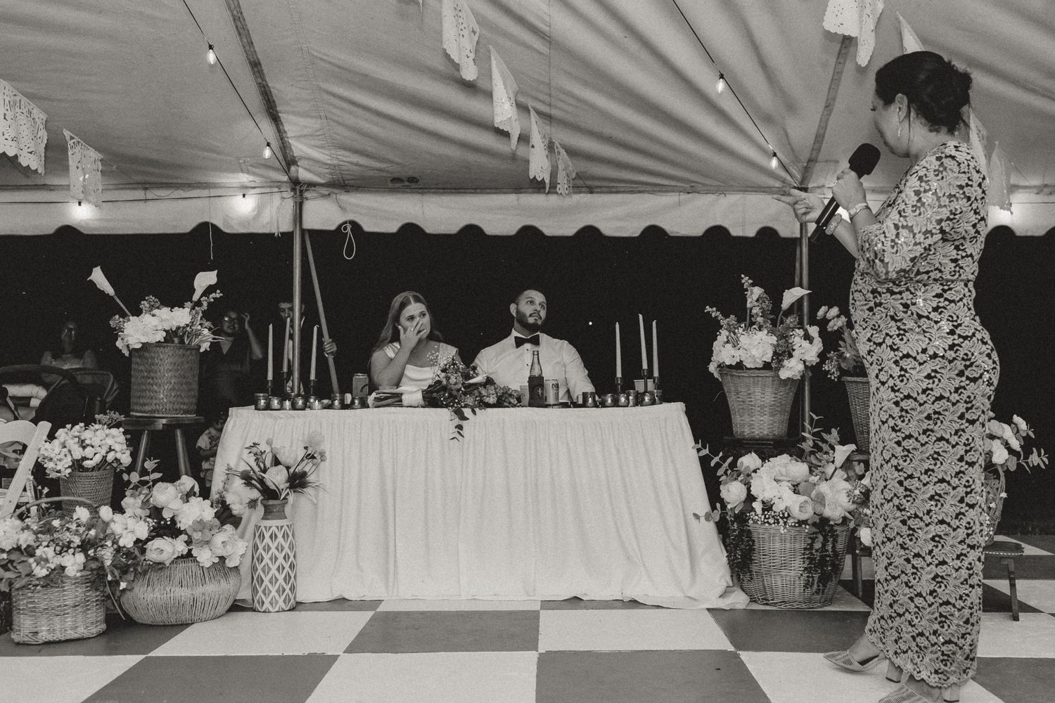 Black and white wedding reception photo with couple at head table, floral arrangements, and guest giving a toast.