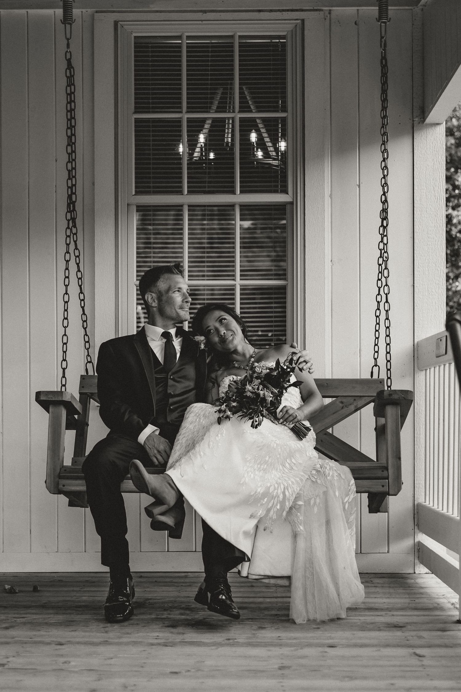Black and white photo of a bride and groom sitting on a porch swing, bride leaning on groom holding bouquet.