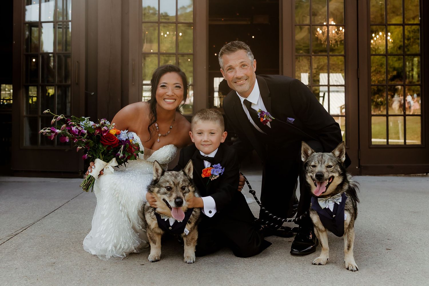 Bride and groom pose with young boy and two dogs on wedding day outside elegant venue doors.