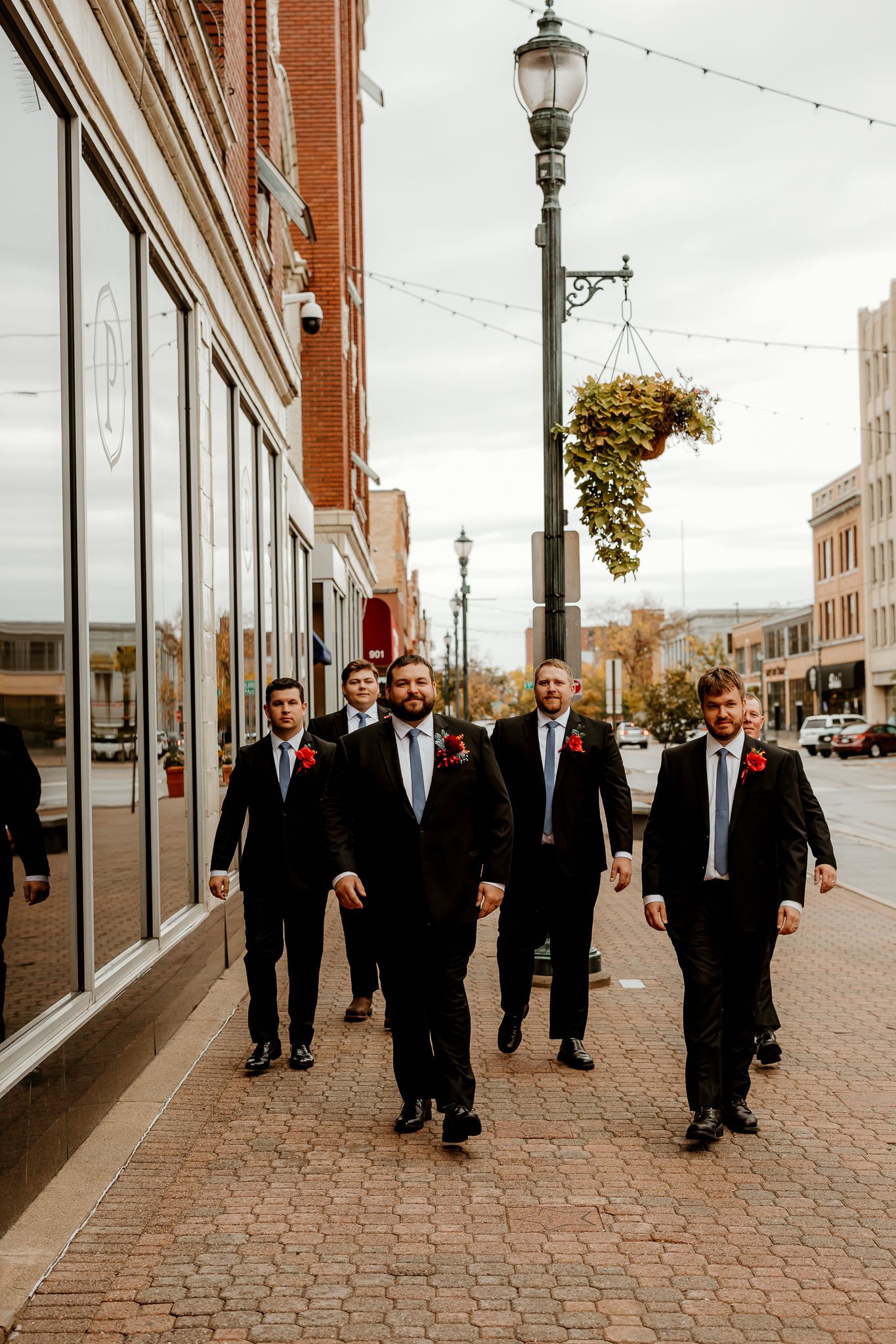 Groomsmen in black suits and red boutonnieres walk together down a brick sidewalk past downtown storefronts on a wedding day.