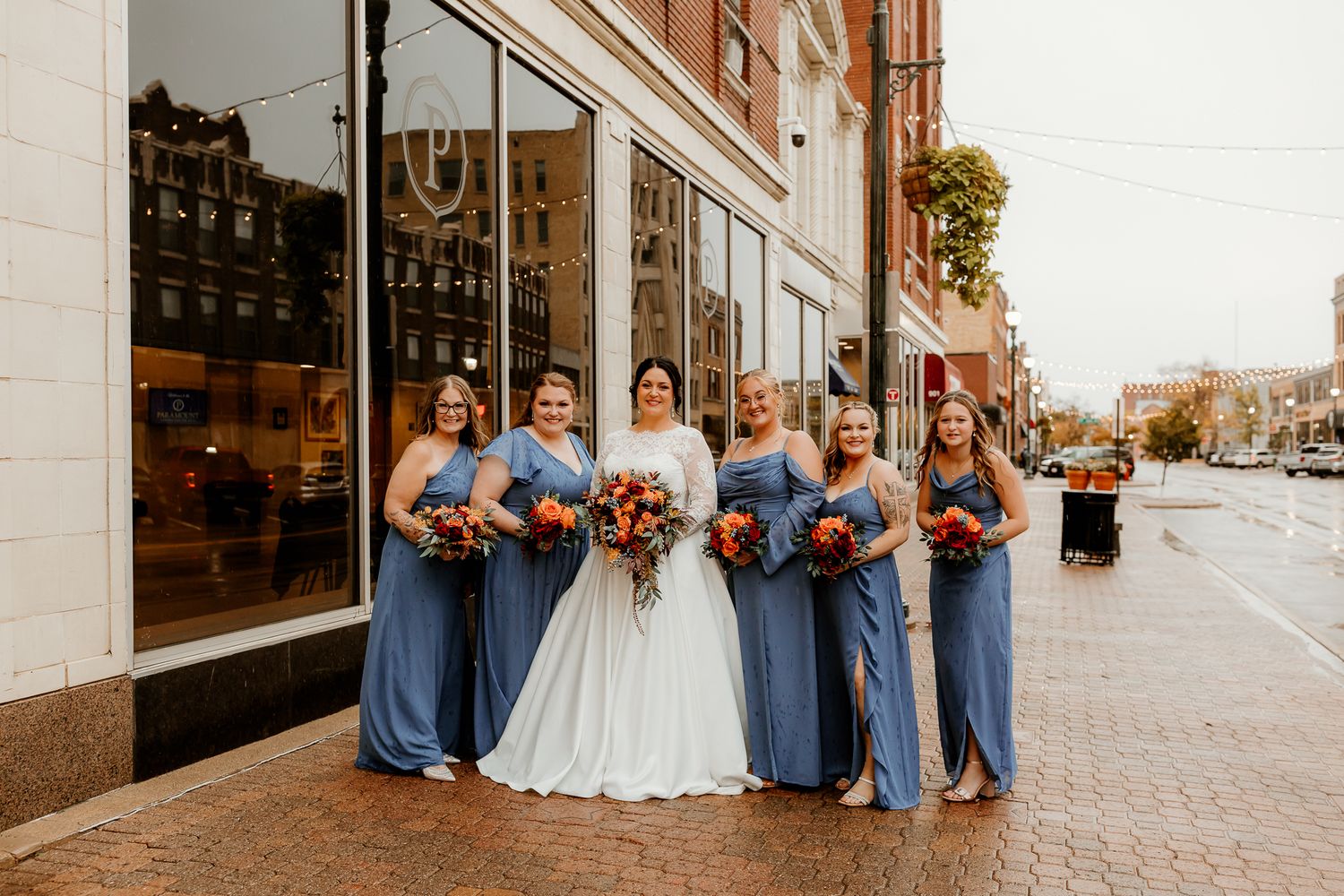 Bride in white gown stands with five bridesmaids in dusty blue dresses holding autumn bouquets on a brick sidewalk outside a city storefront