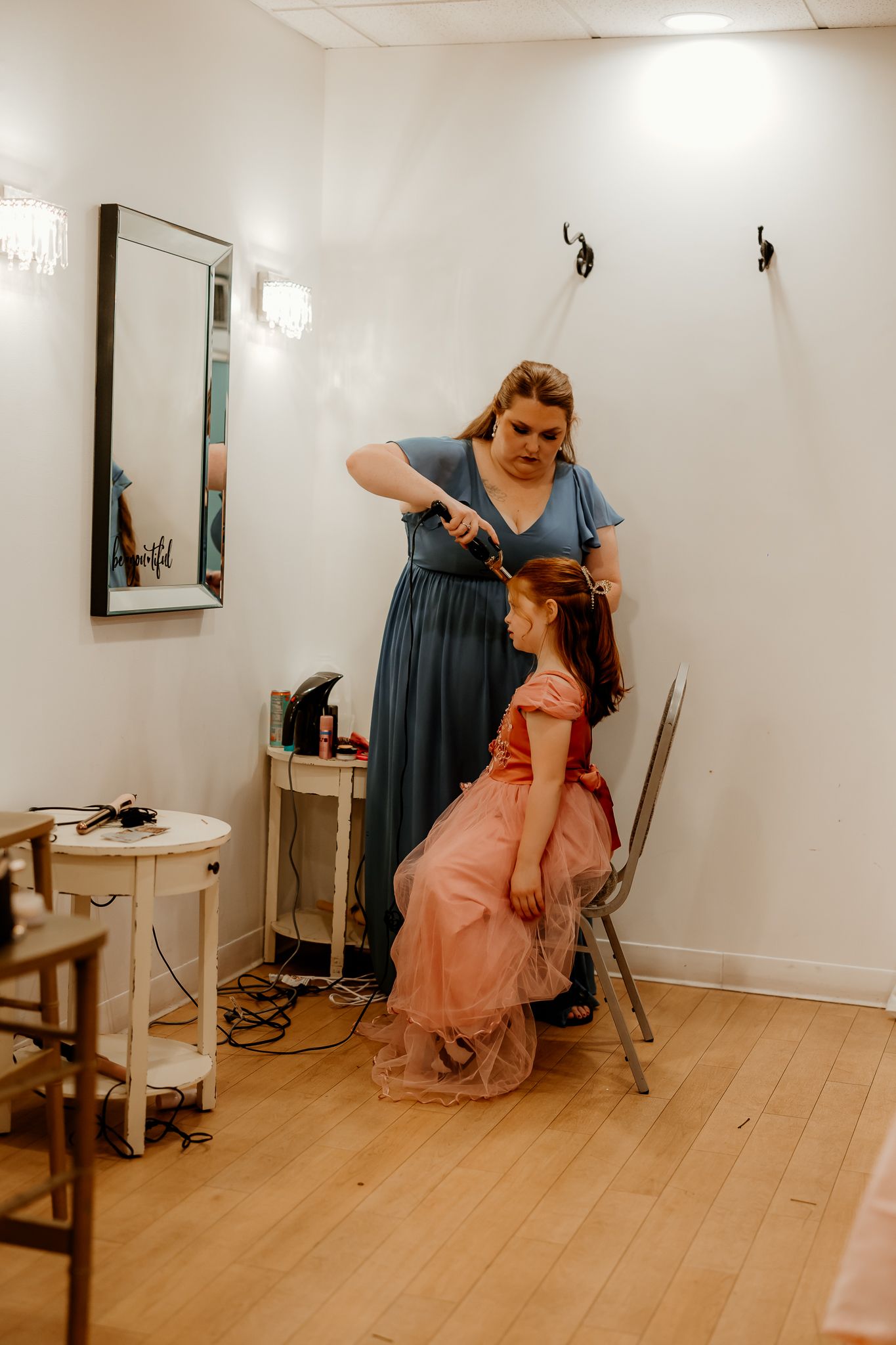 A woman in a blue dress curls the hair of a young girl in a pink tulle dress seated on a chair in a bright, mirrored dressing room.