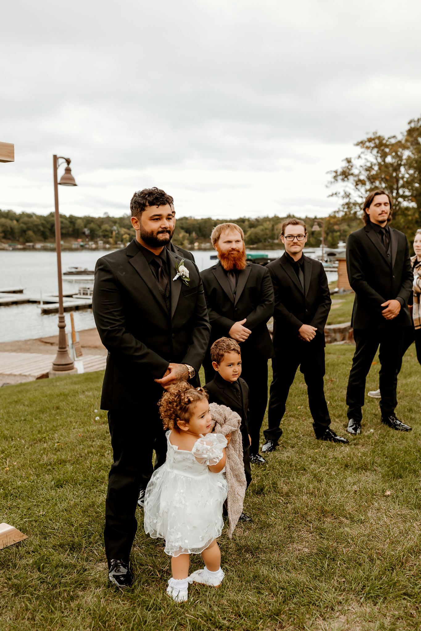 Groomsmen in black suits stand on a lakeside lawn at an outdoor wedding while a flower girl in a white dress and a young ring bearer wait.