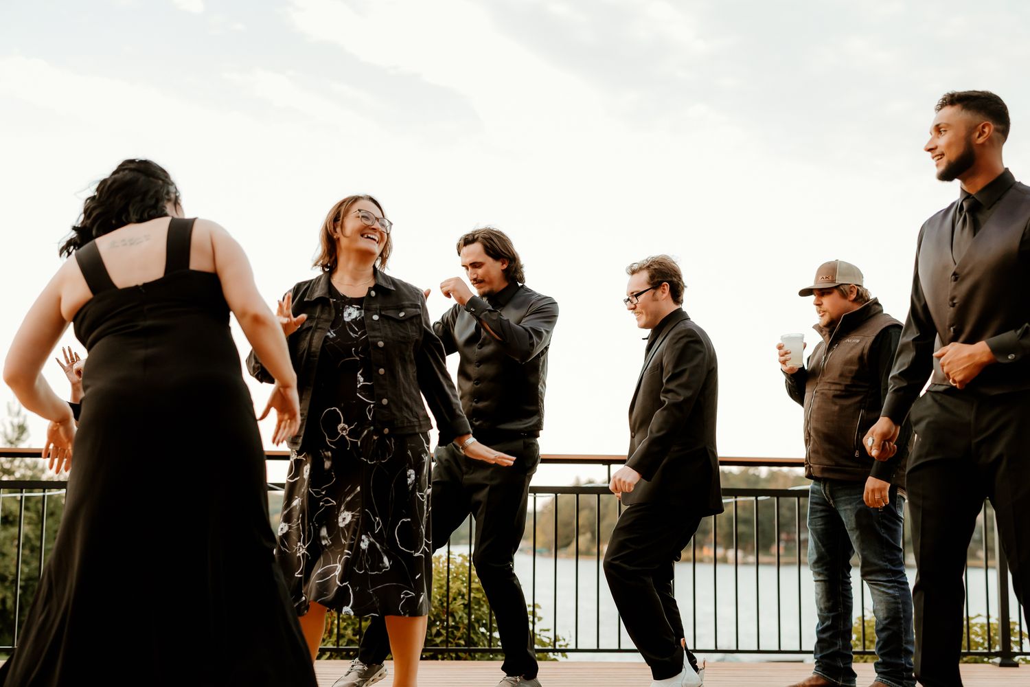 Six friends in dark, stylish outfits laugh and dance together on an outdoor terrace overlooking a scenic waterfront on a bright day.