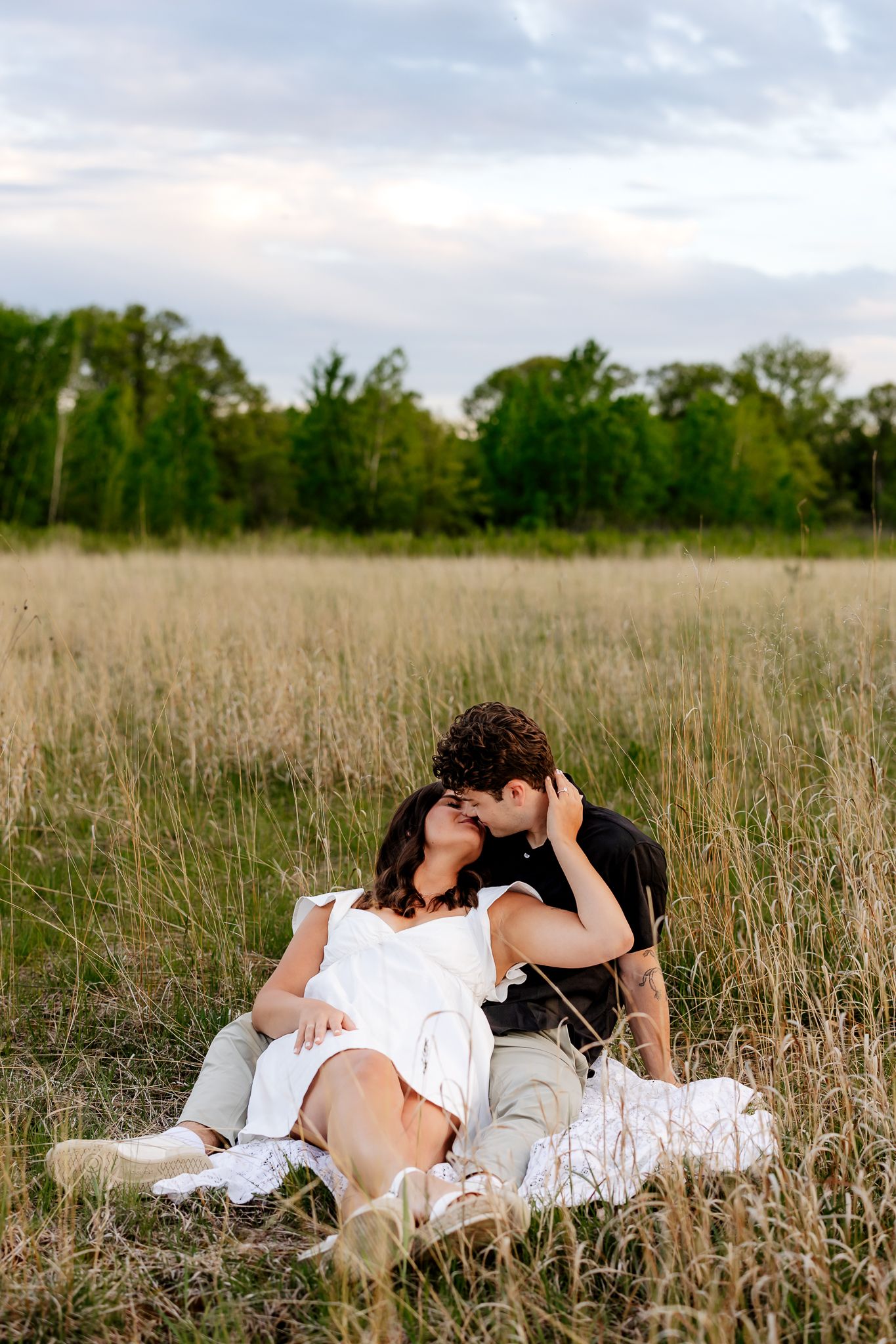 Couple sharing a kiss while relaxing on a blanket in a tall grass field at sunset.
