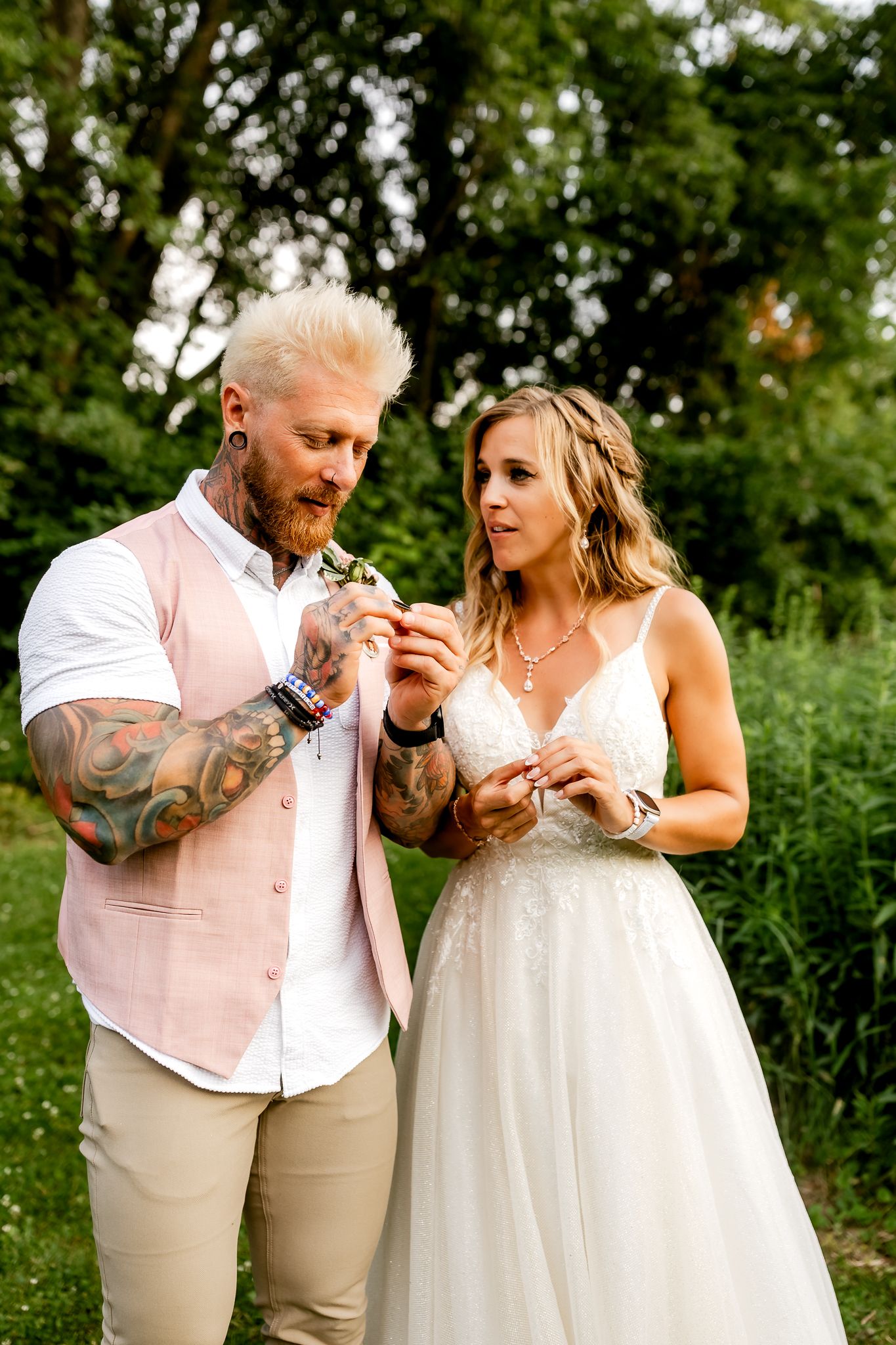 Tattooed groom in pink vest and bride in lace wedding dress exchanging rings in a lush green garden.