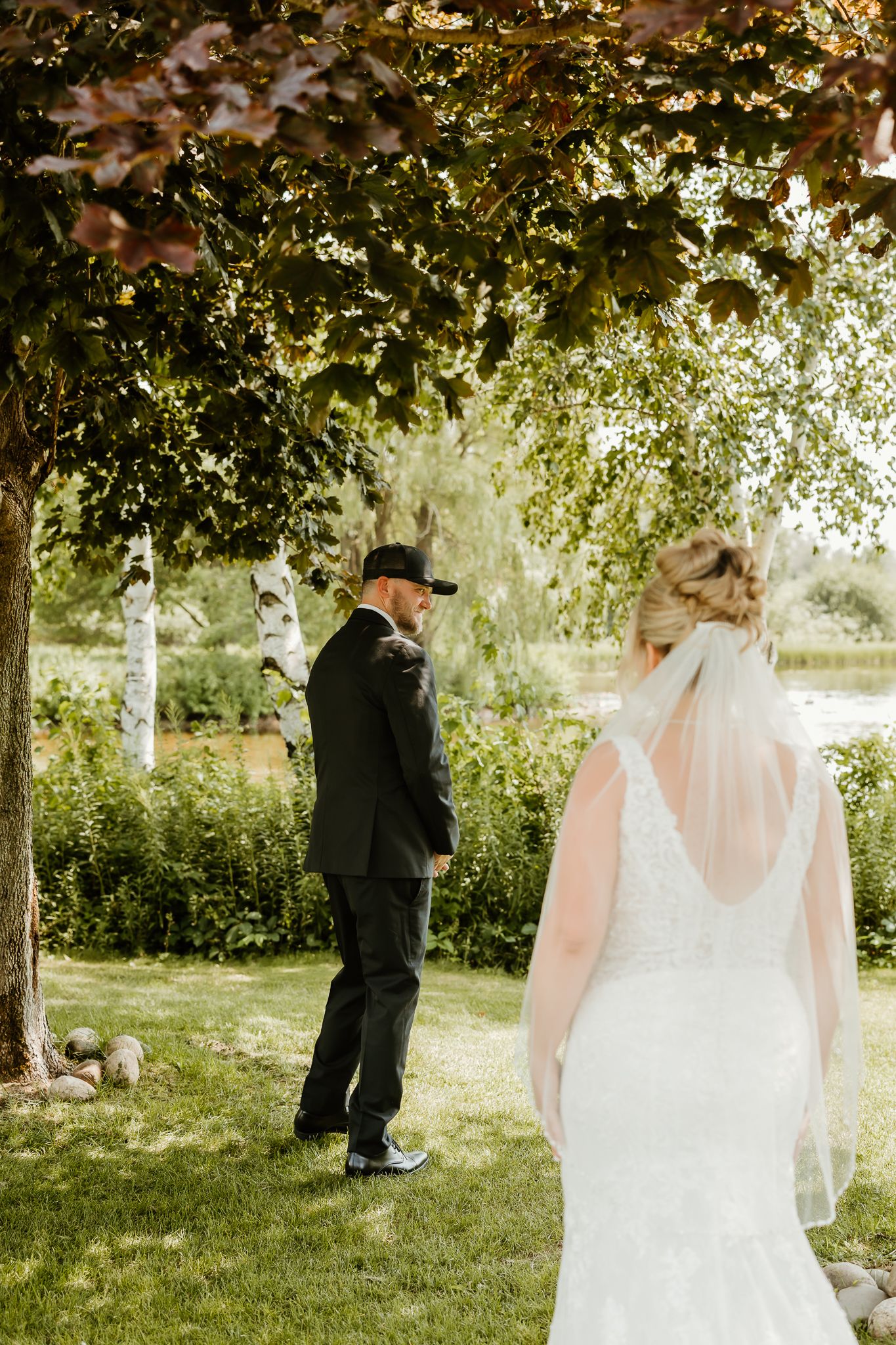Bride in lace gown with veil approaches groom in black suit for a first look moment outdoors near a pond.