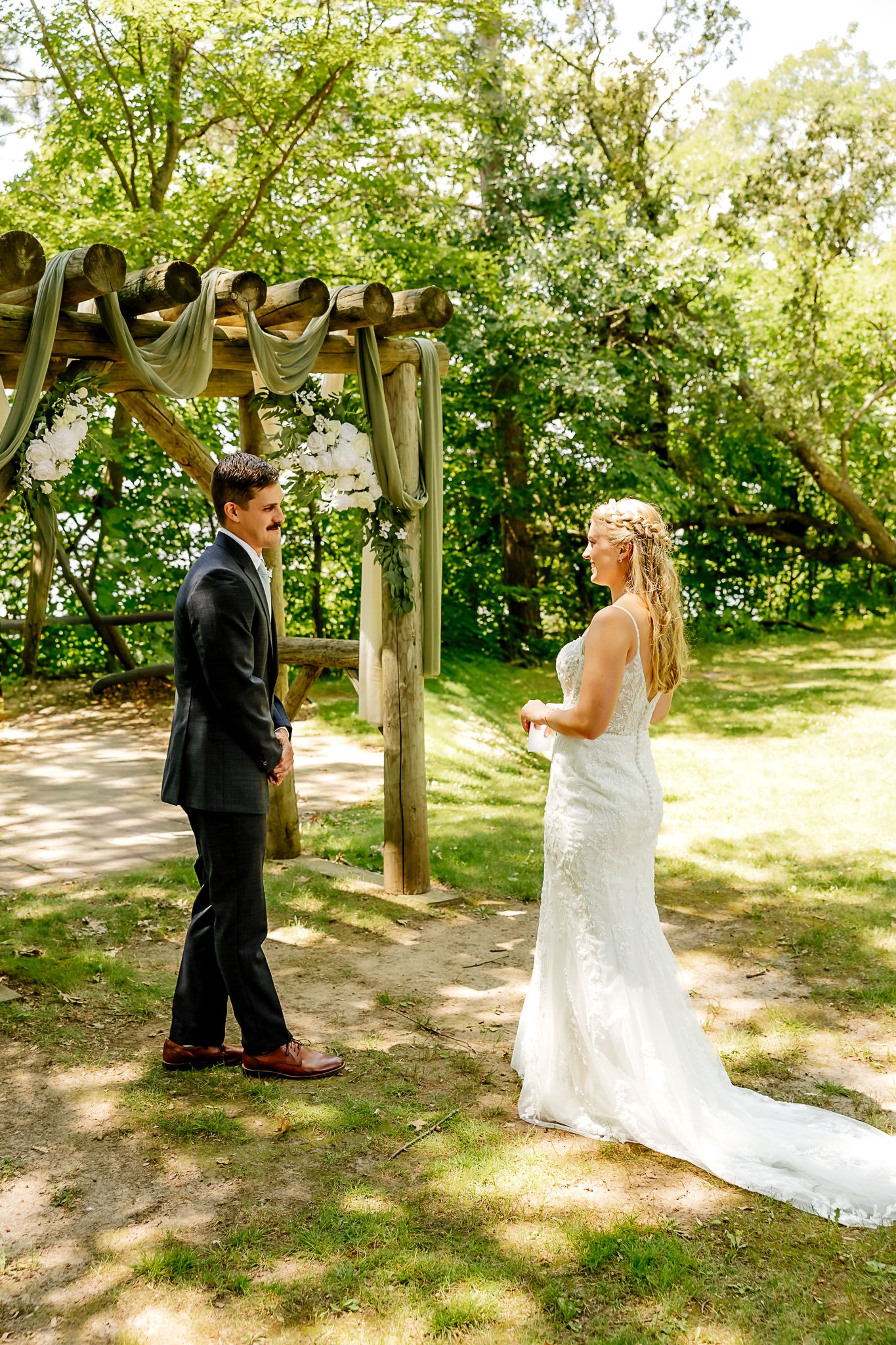 Bride and groom share a first look moment outdoors near a rustic wooden arch surrounded by lush green trees.