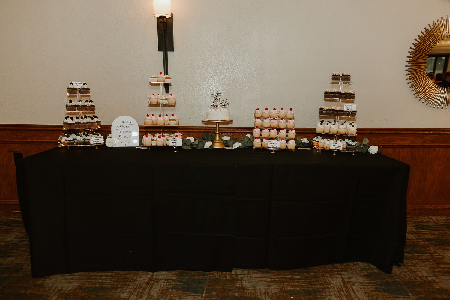 Elegant wedding dessert table with black tablecloth displaying tiered cupcakes, mini cakes, and a small wedding cake centerpiece.
