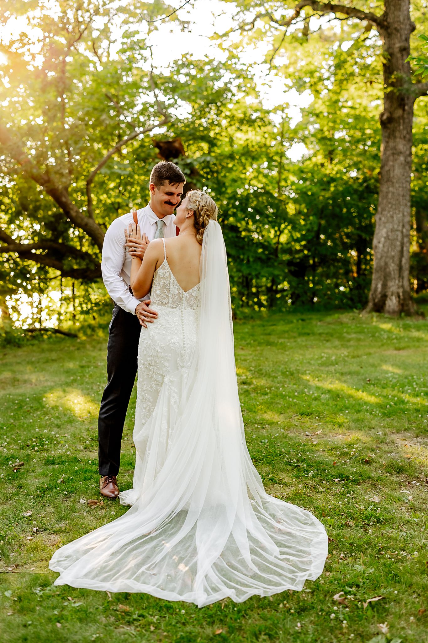 A bride in a lace gown with long veil and groom in suspenders share a romantic moment outdoors at golden hour.