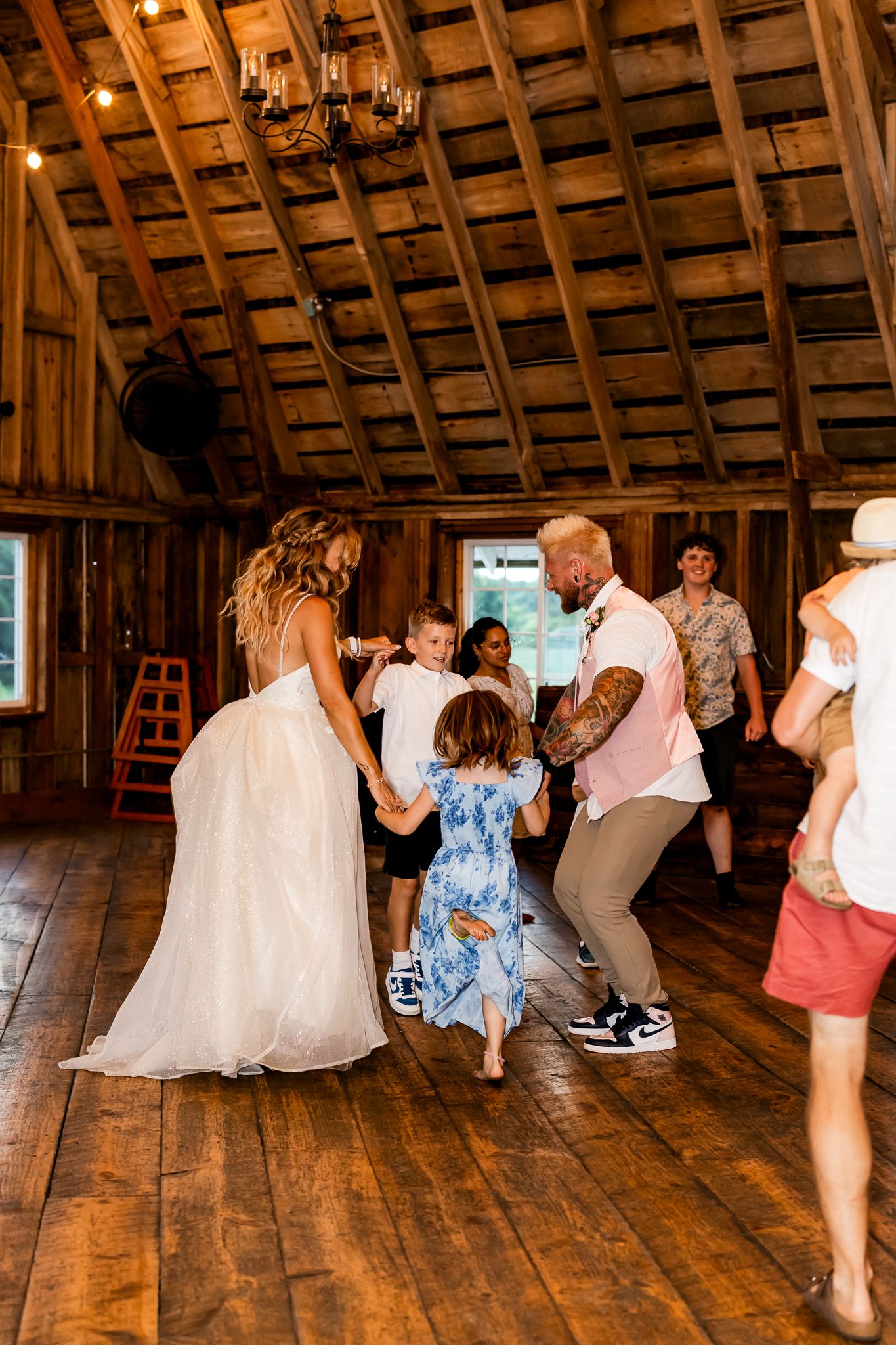 Bride in white gown and guests dancing joyfully with children inside a rustic wooden barn wedding reception.