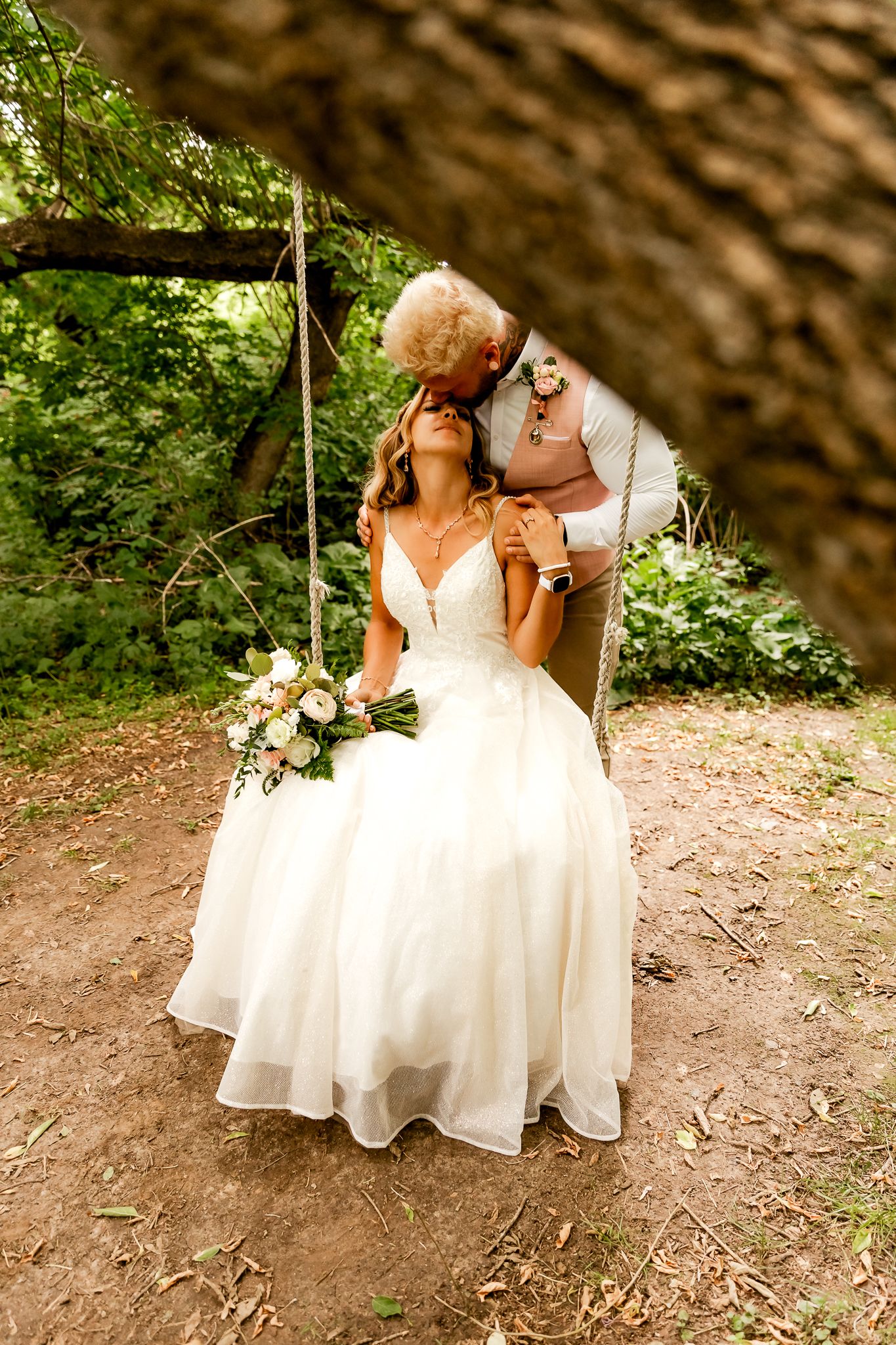 Bride in white gown sits on swing as partner kisses her cheek in lush green outdoor wedding setting.