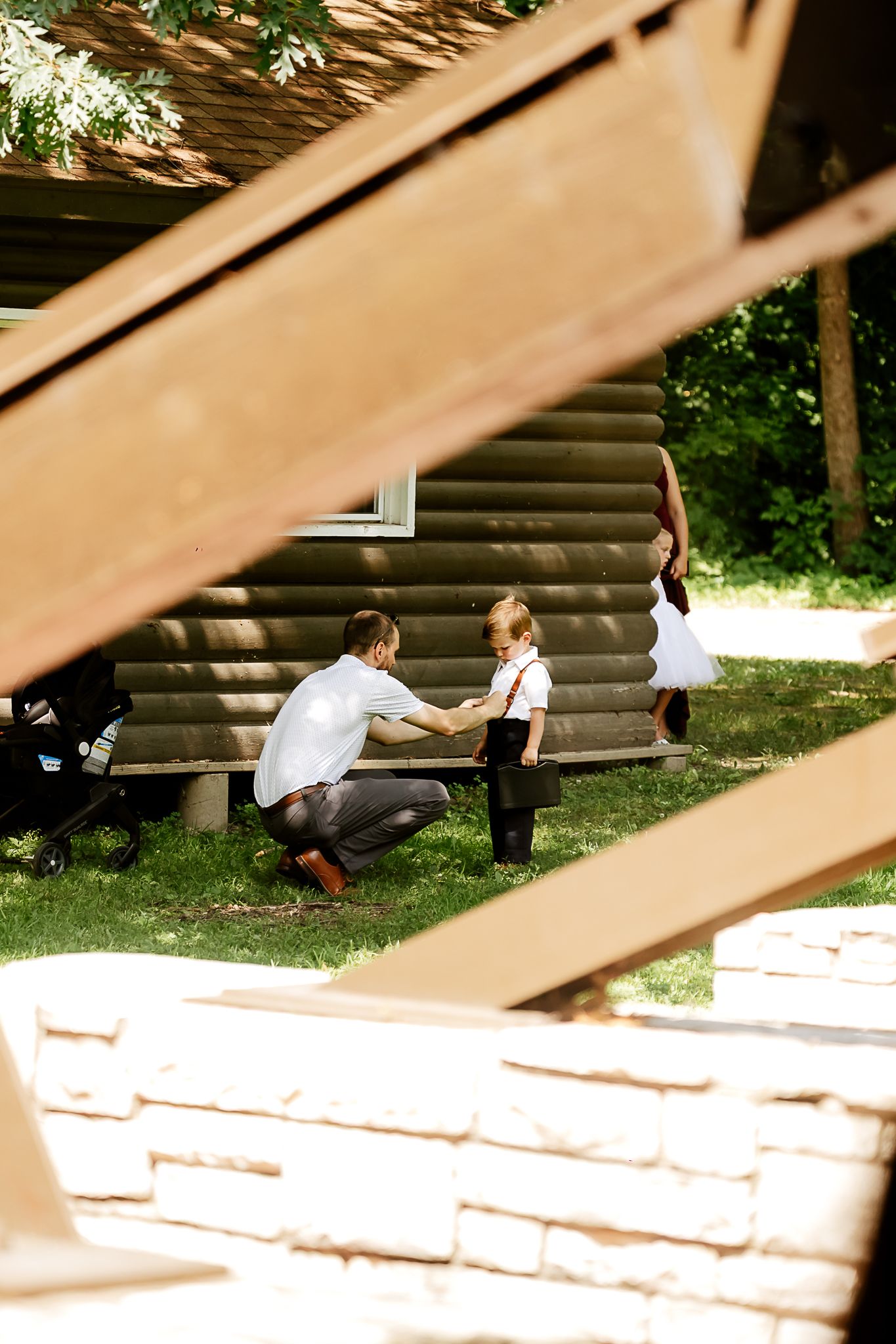 A father kneels before his young children near a wooden cabin bench surrounded by lush green trees outdoors.