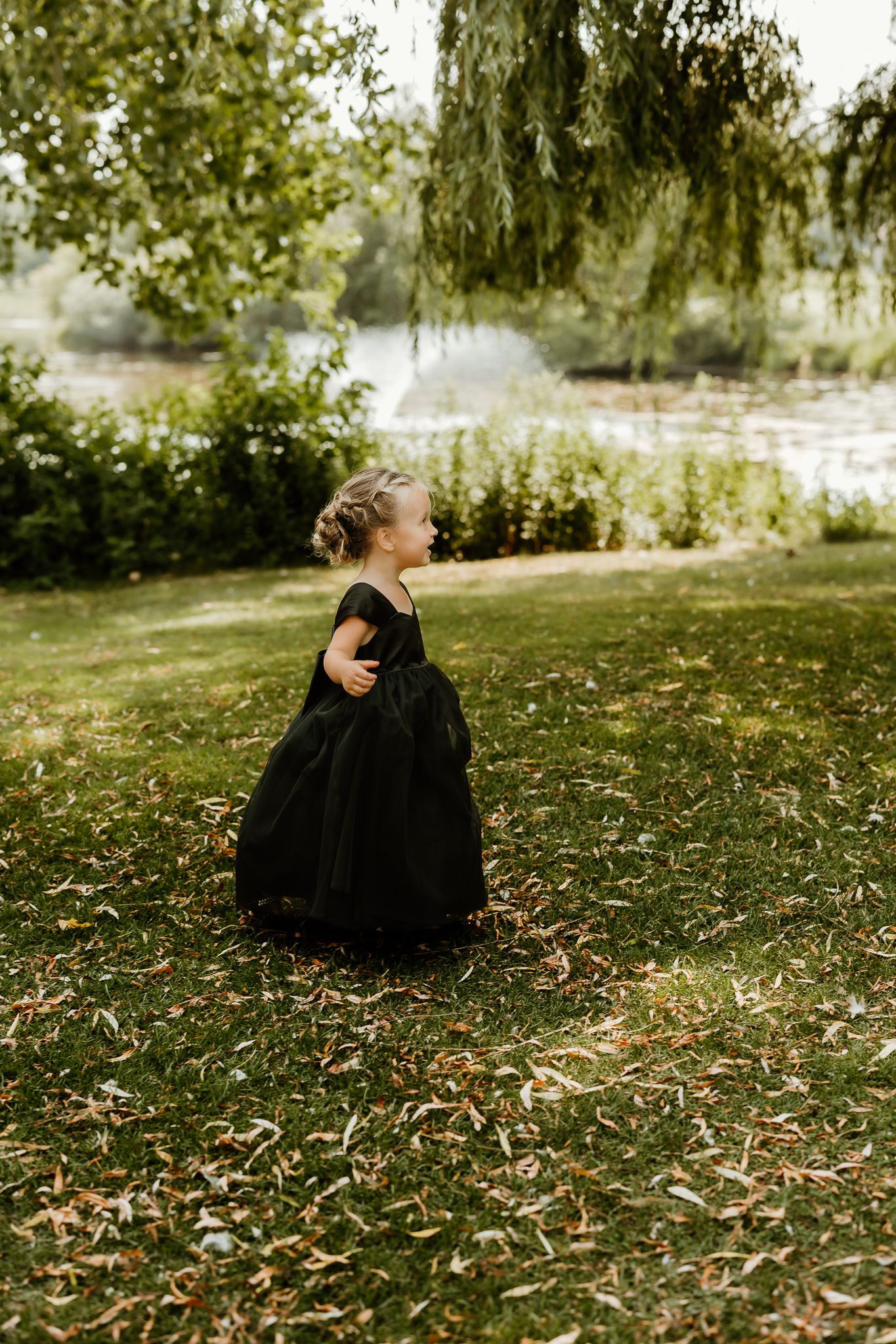 A young girl in a black ball gown dress plays outdoors near a pond surrounded by lush green trees.