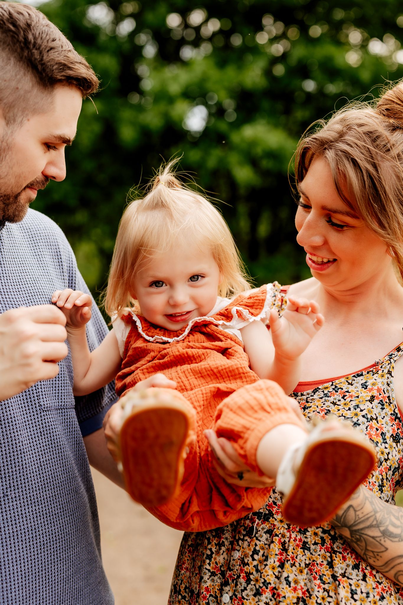 Smiling parents admiring their cute red-dressed toddler outdoors in nature.