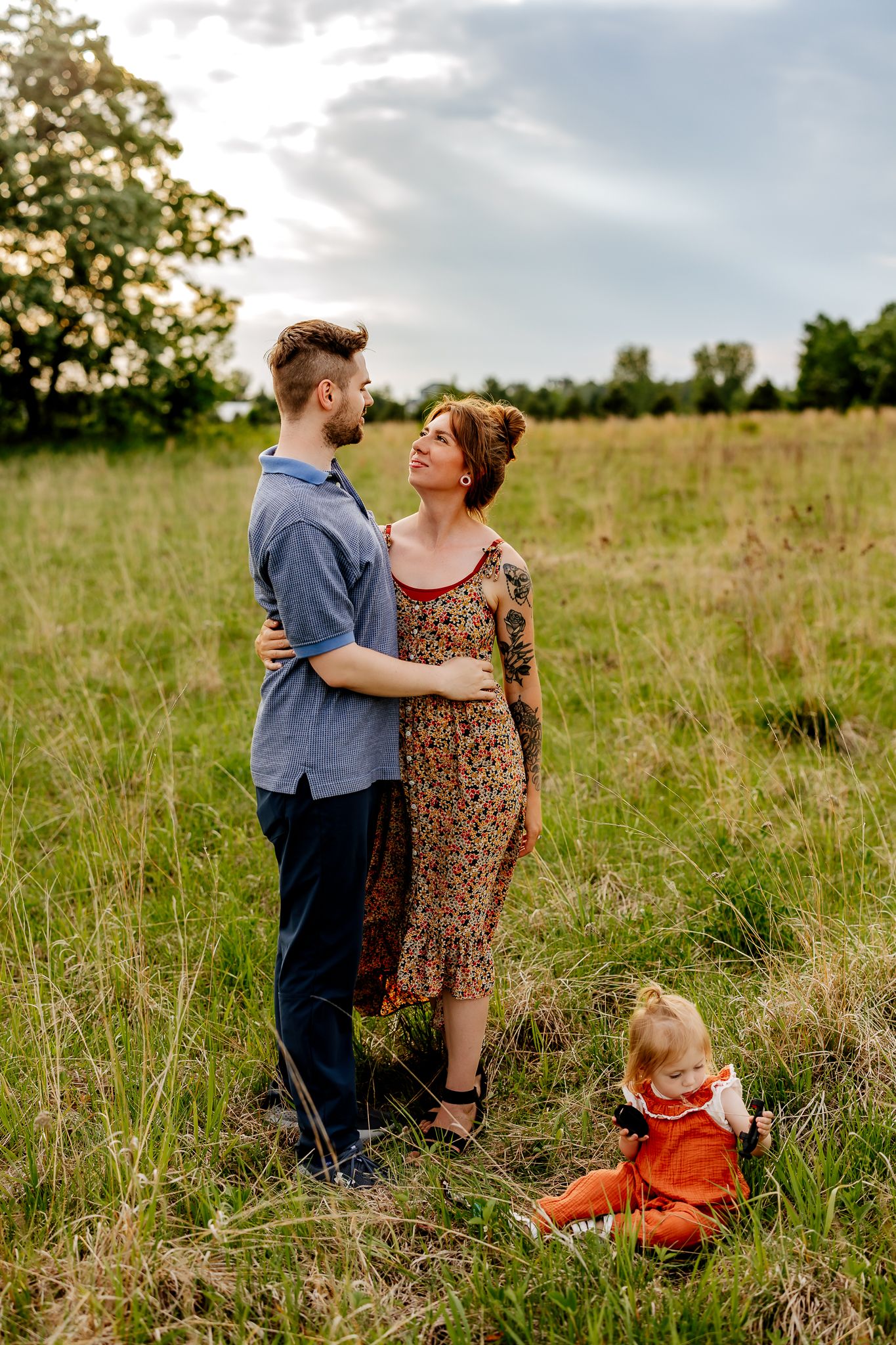 Young family with toddler poses in golden hour meadow, parents embracing lovingly.