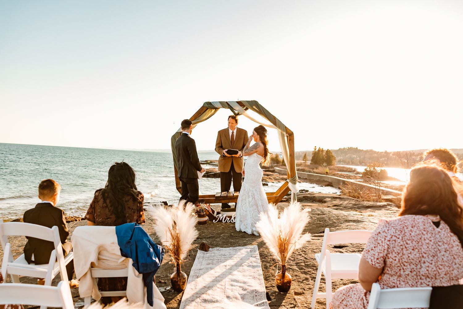 Couple exchanging vows at a romantic beach wedding ceremony at sunset with seated guests.