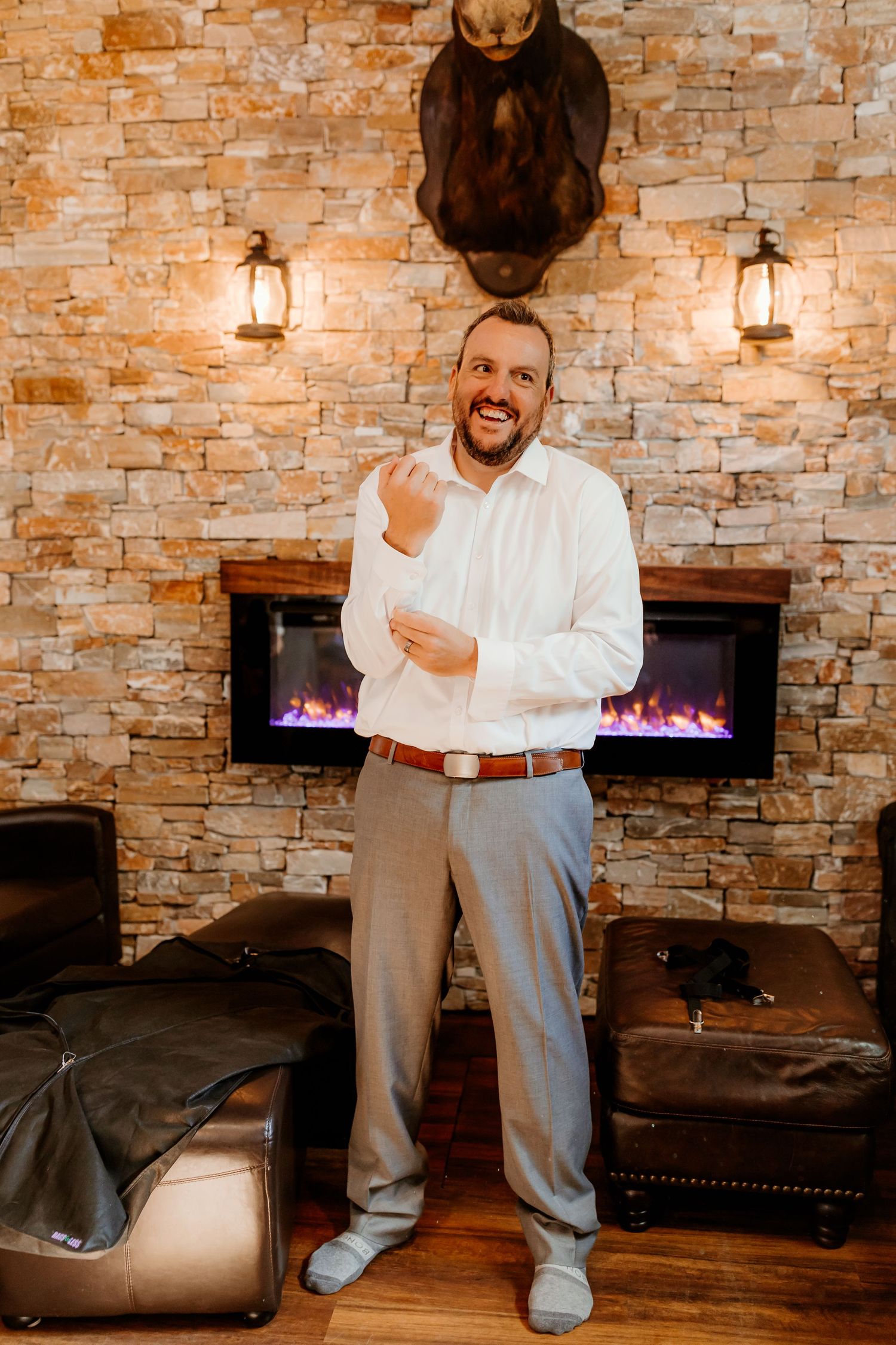 Smiling man in white dress shirt and gray pants adjusts his cuff in front of a stone wall with a lit modern fireplace.